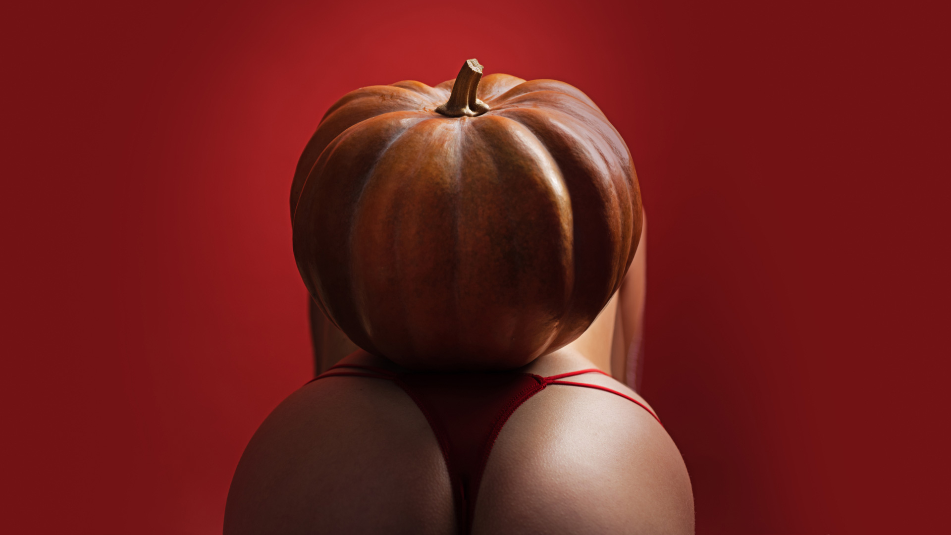Ass of a woman wearing revealing, red thong bending over with orange pumpkin on her back.