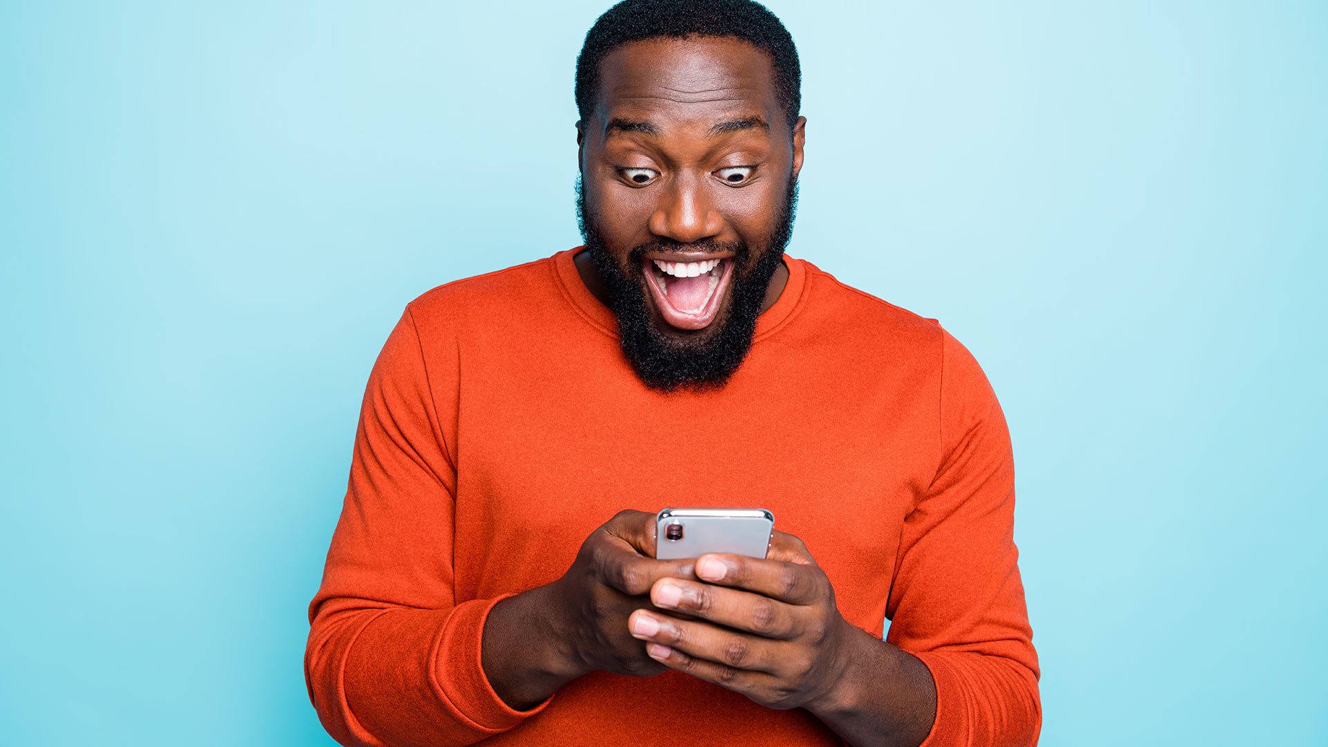 Brunette man in orange long-sleeve shirt, holding his phone with both hands and looking at it excited. Light blue background.