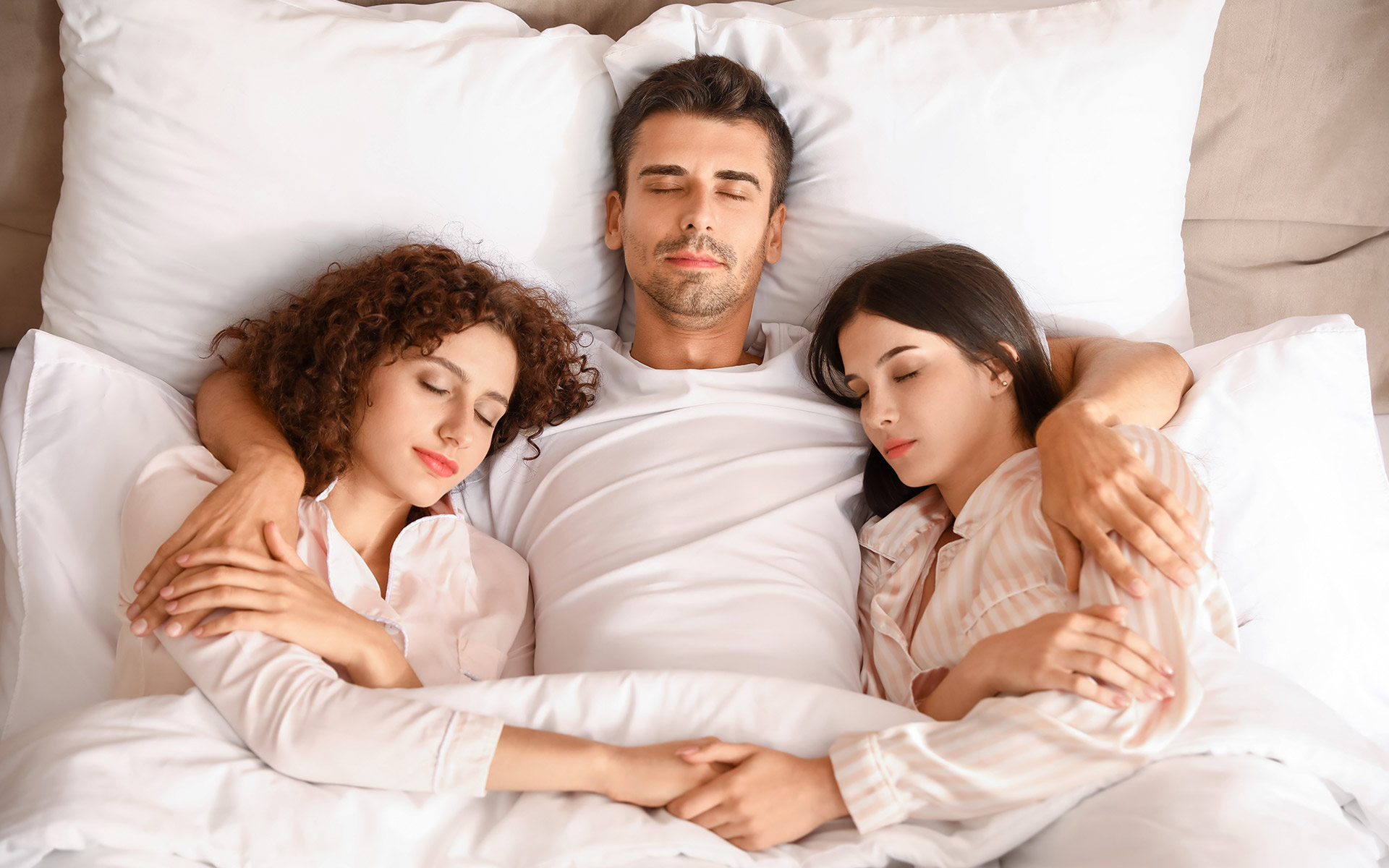 Sleeping man lying in bed hugging two sleeping women. His arms are around each woman and they are holding each other's hand.