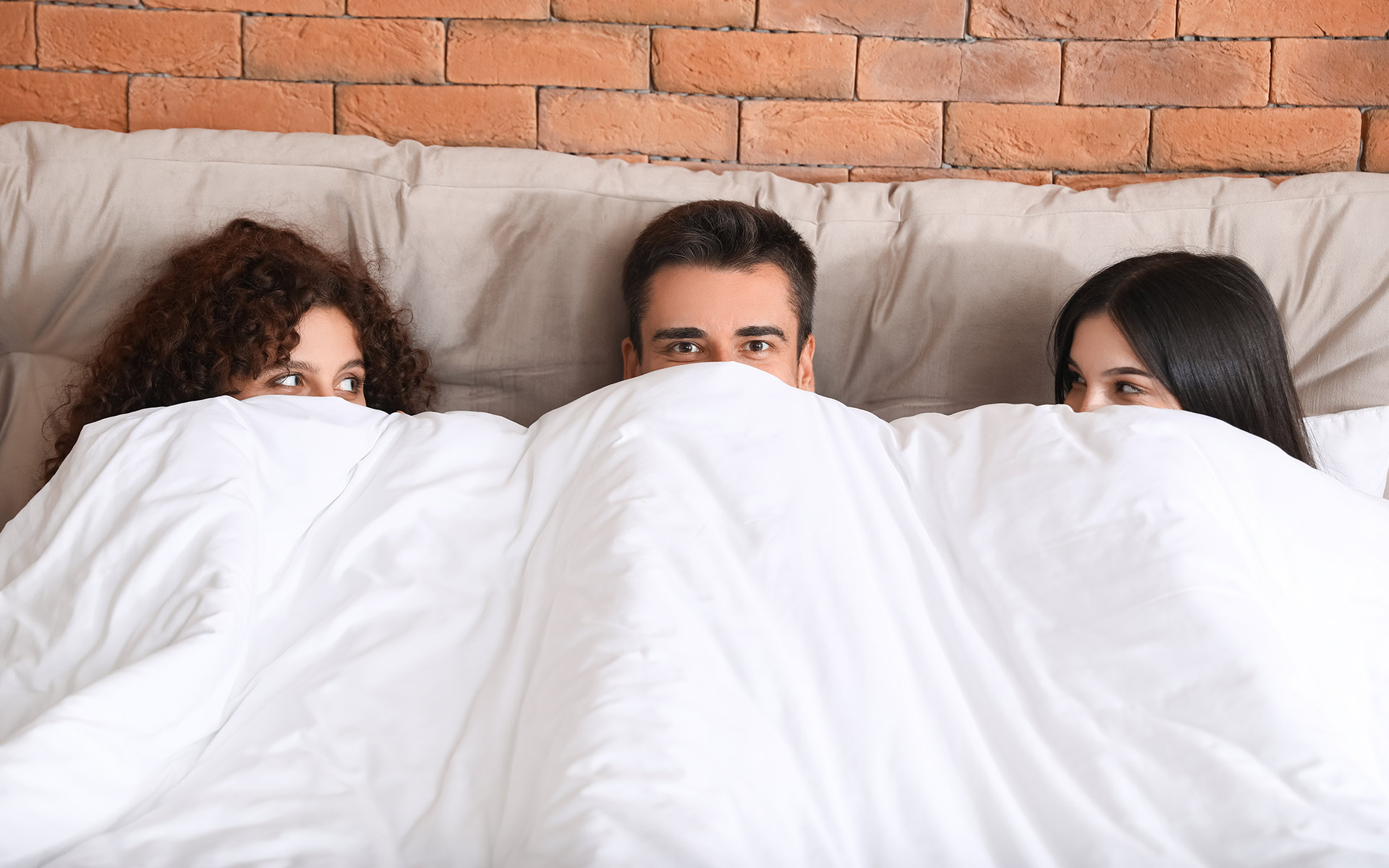 Brunette man in bed between two brunette women, all covering half their faces with the duvet and looking at each other.