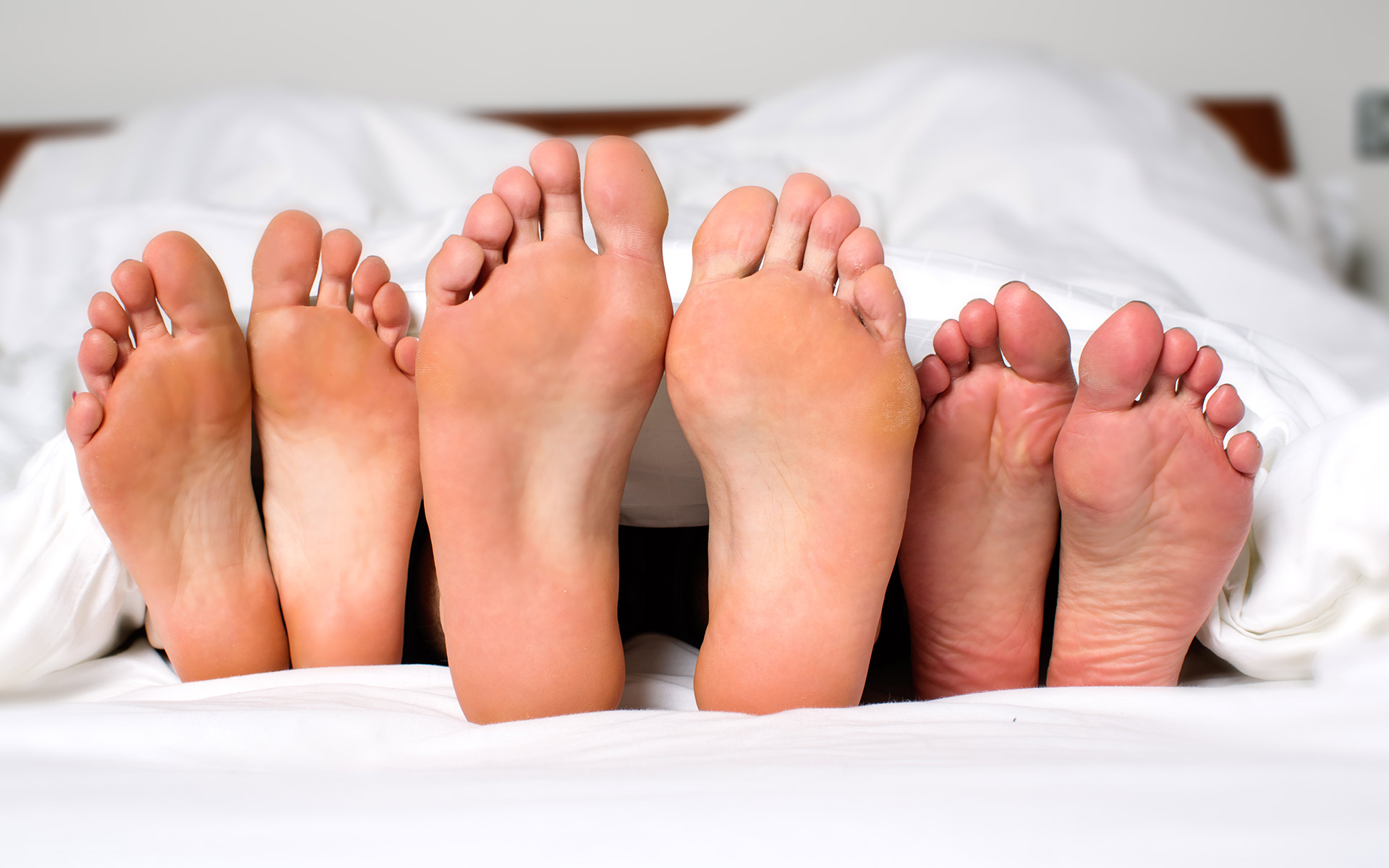 Three pairs of feet lying in bed, coming out of a white duvet. The middle pair of feet is larger than the other two.