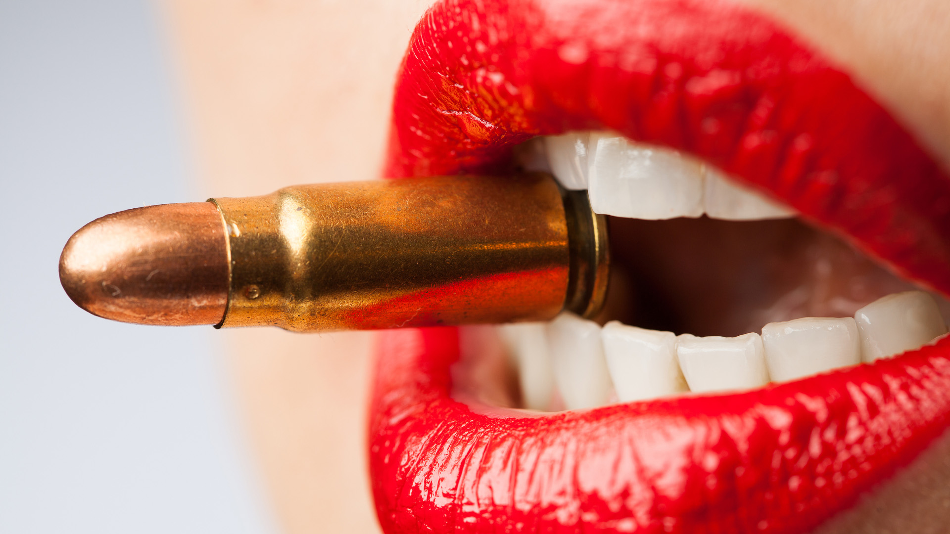 Mouth of a woman, parted red lips biting down on a golden bullet.