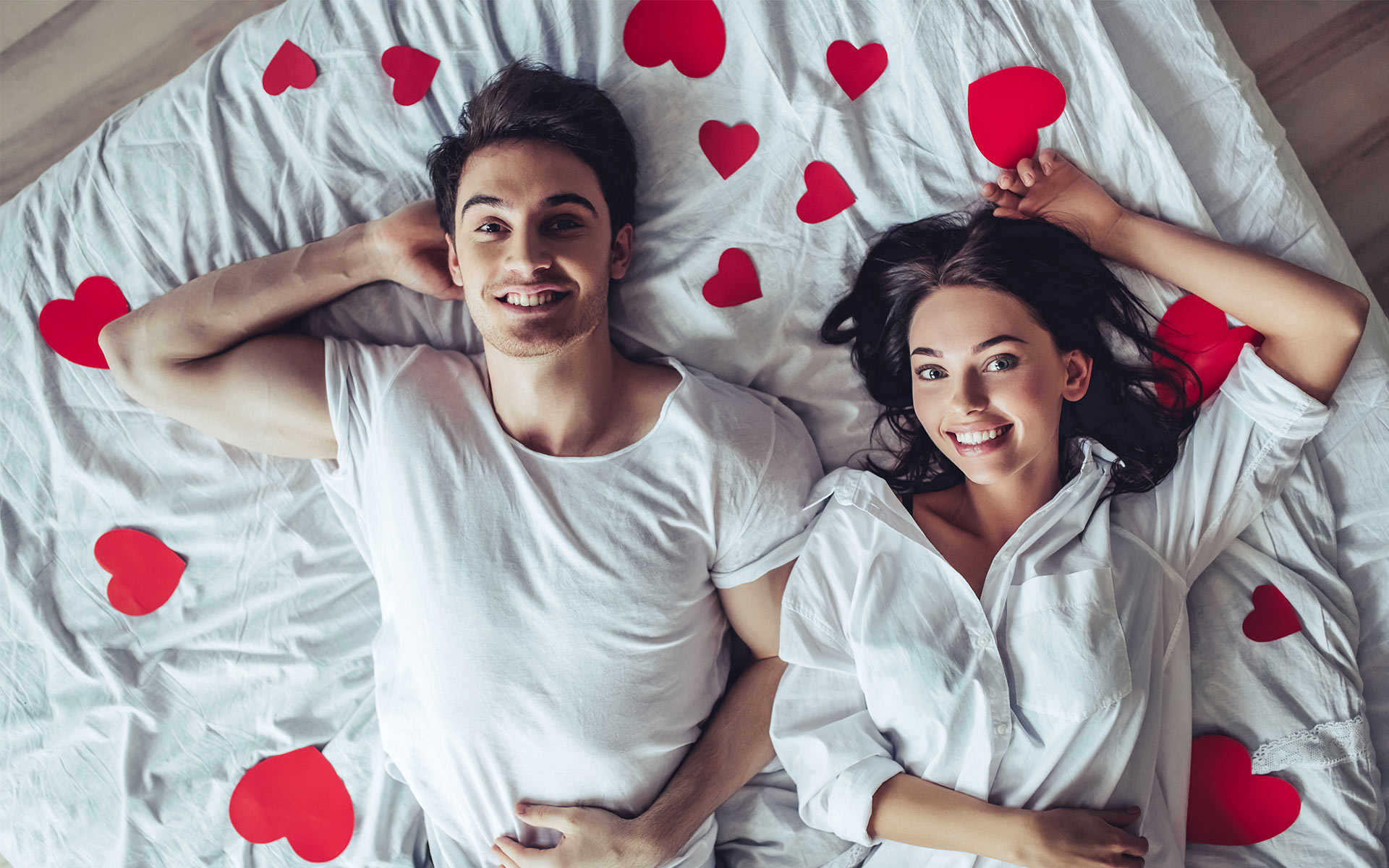 Brunette man and woman wearing white shirts lying next to each other in bed with red love-hearts around them, smiling.