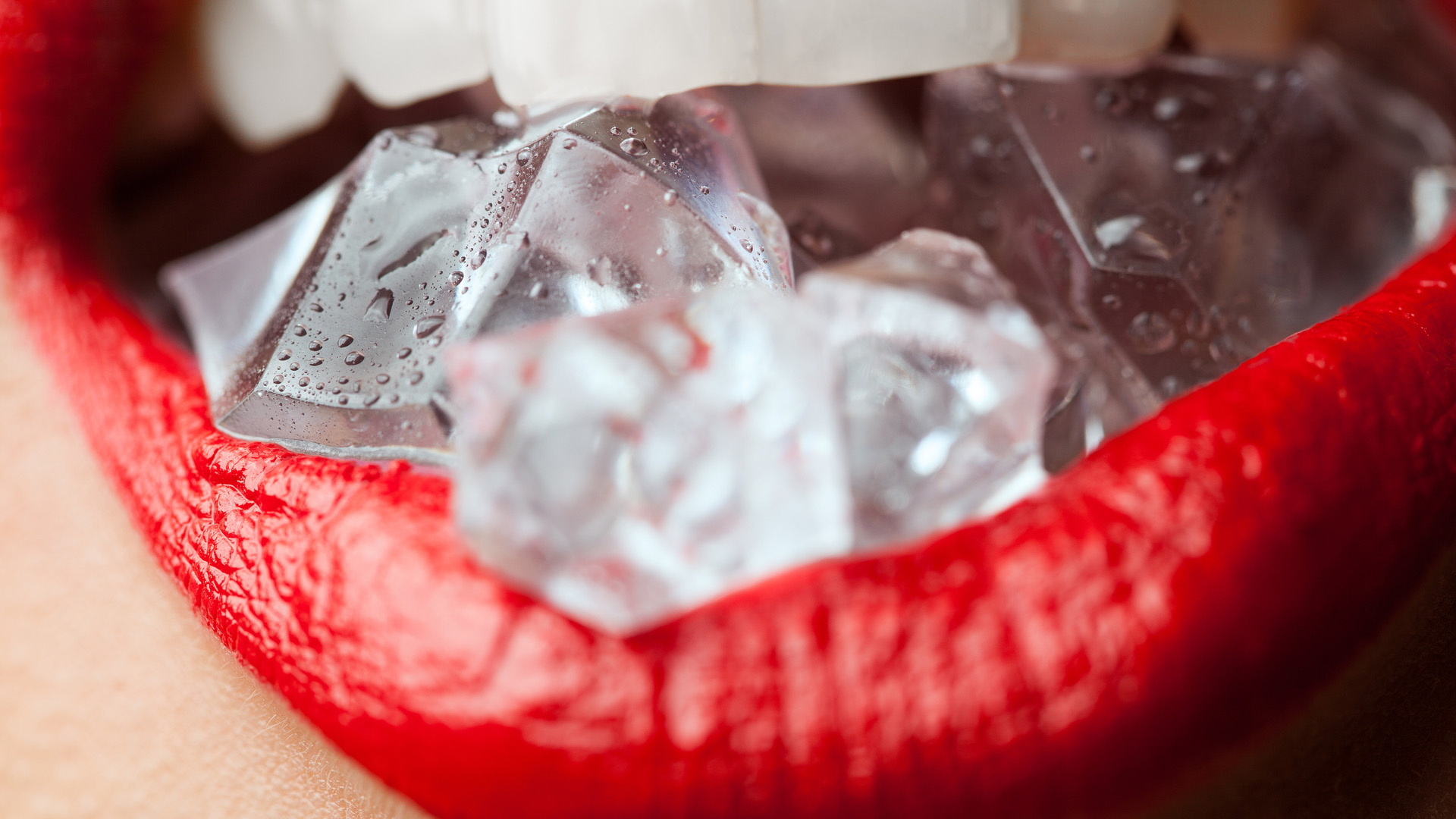 Open mouth of a woman in red lipstick biting down on ice cubes.