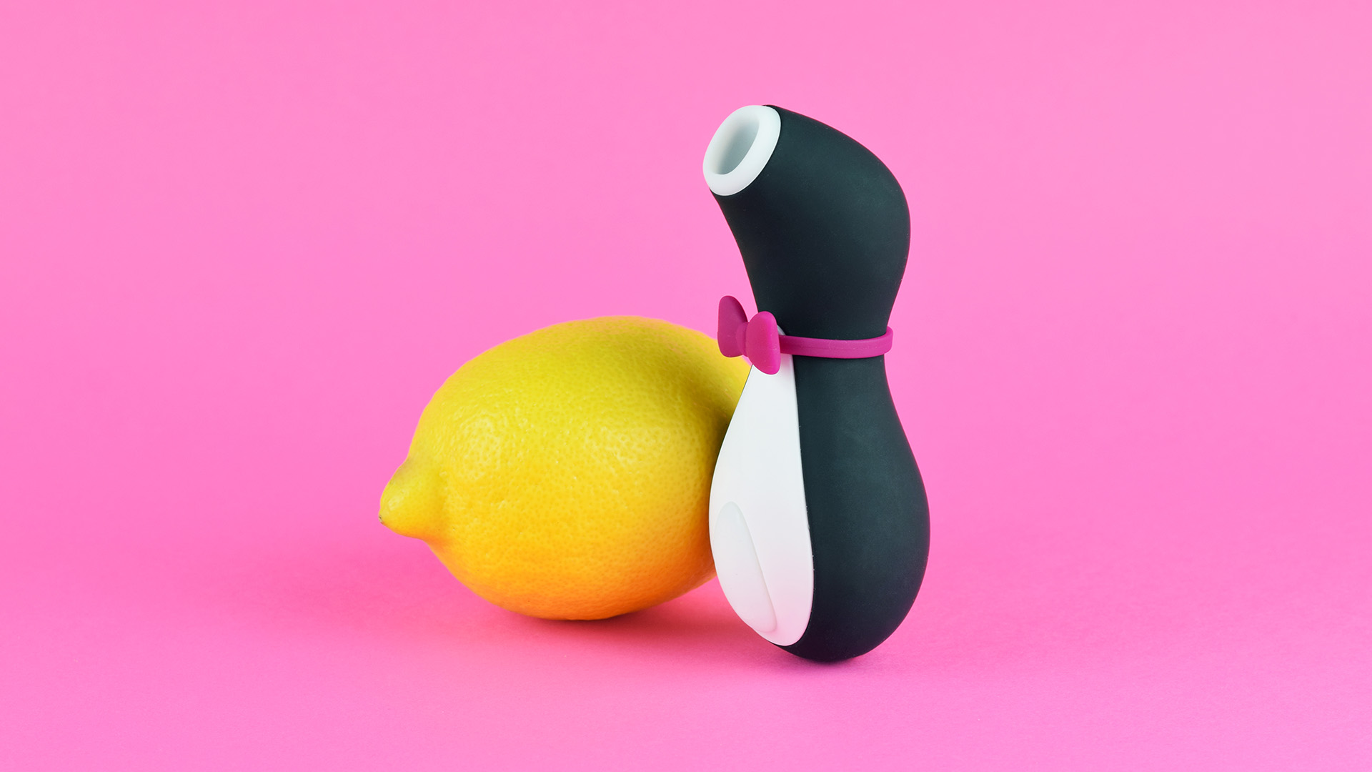 Black vibrator with white tip and button, pink bow around it, next to yellow lemon in front of pink background.