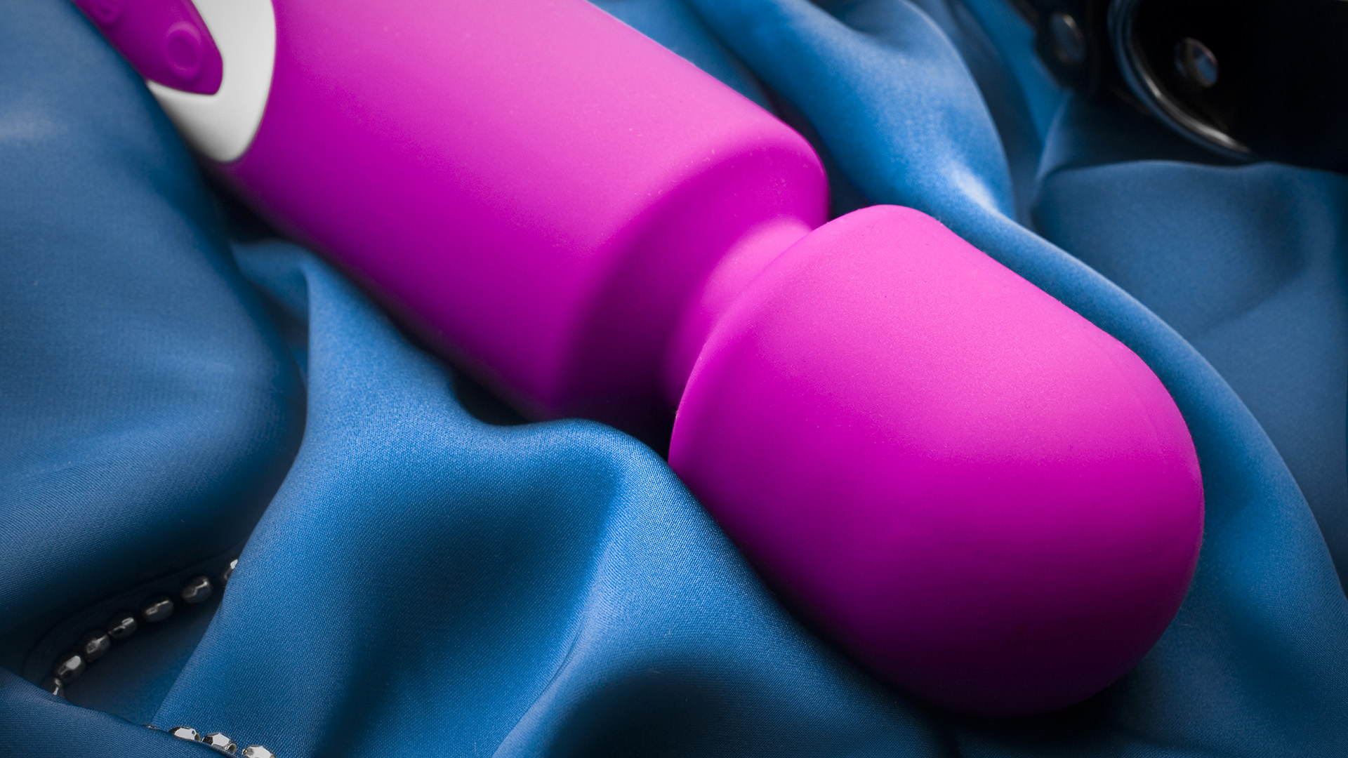 Pink wand vibrator with white border, on blue silky sheets next to chains and chokers.