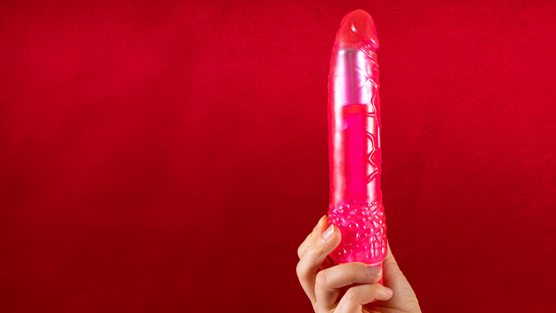 Hand holding a nearly see-through vibrator with pink hue, in front of dark red background.
