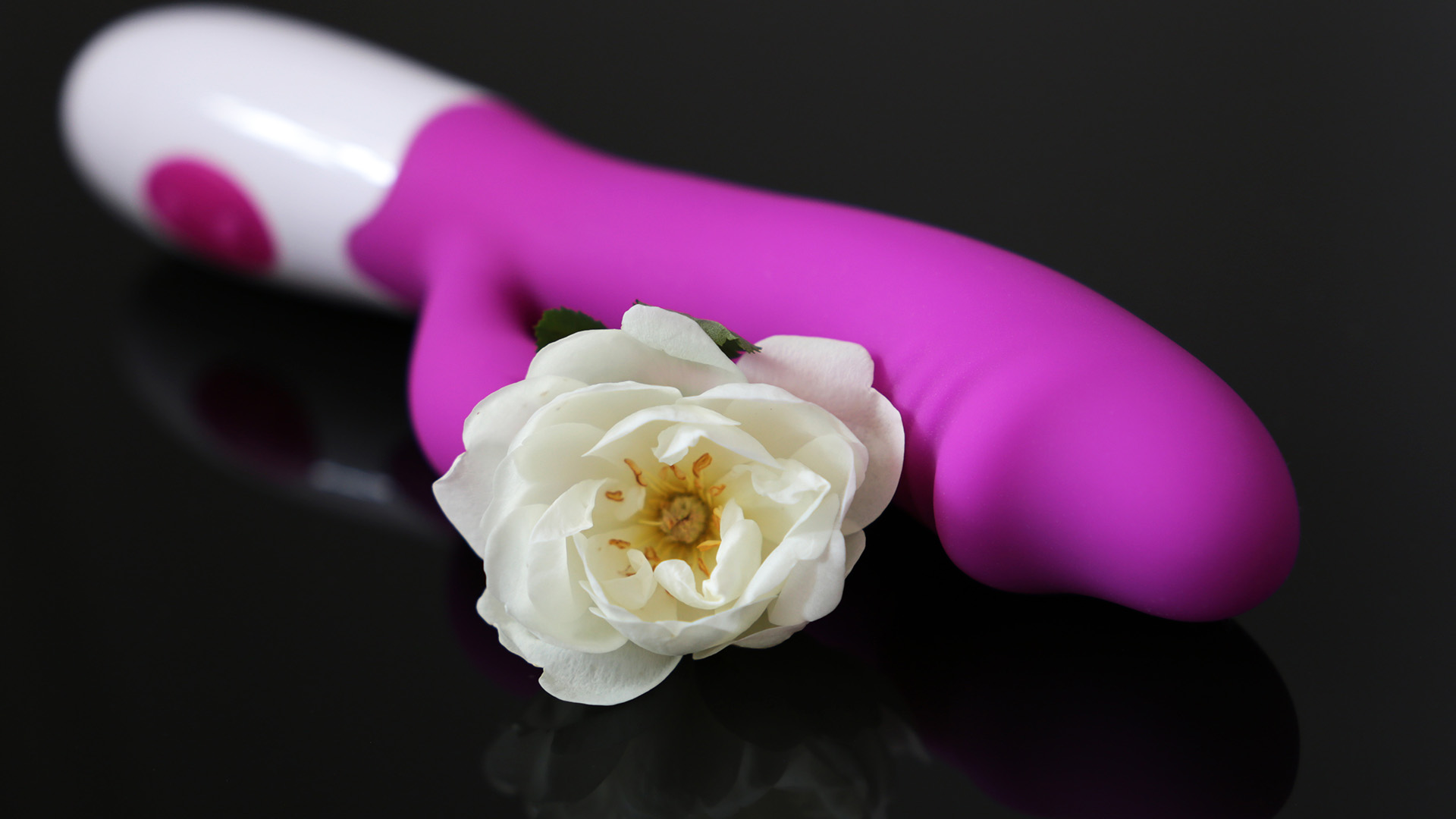 Pink g spot vibrator with white handle next to beige flower, both on a black reflective surface.