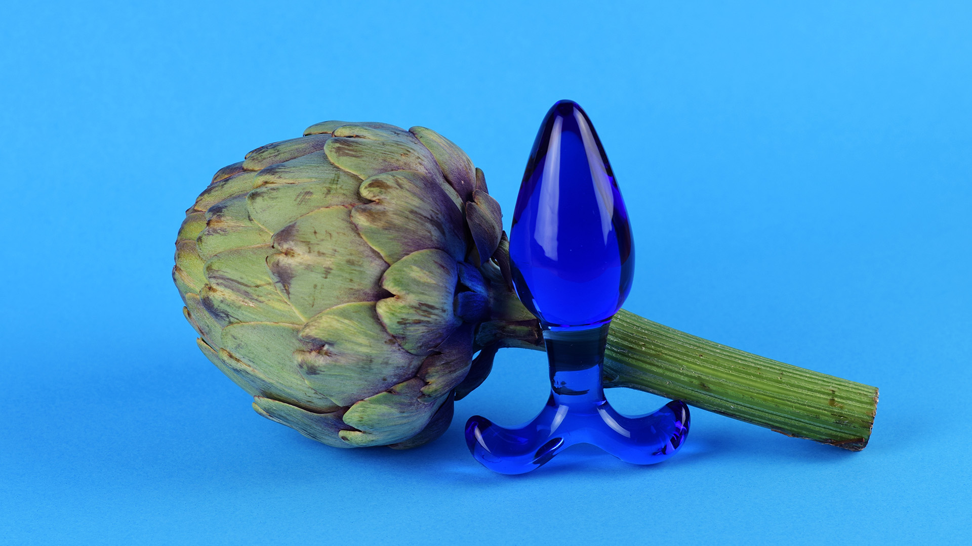 Dark blue see-through butt plug in front of artichoke. Light blue background.