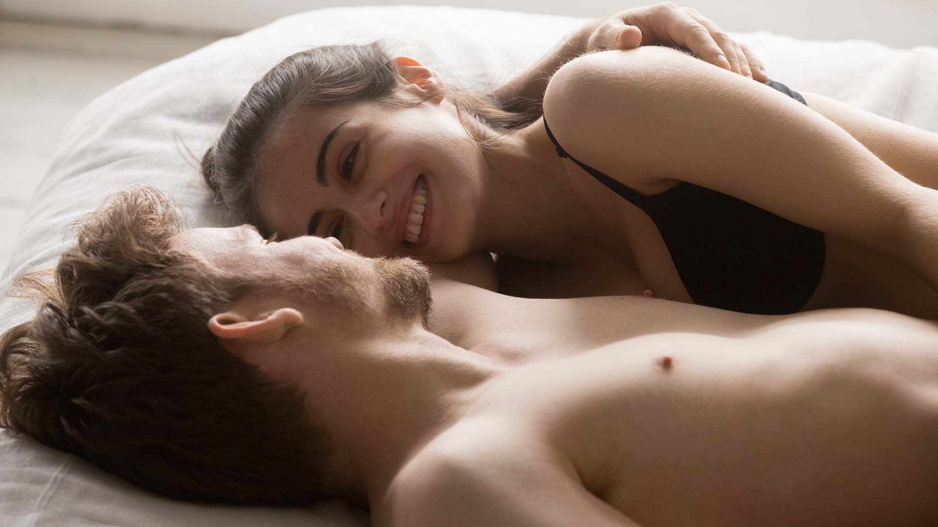 Brunette man and woman lying in bed together and smiling while conversing in their underwear. White sheets.