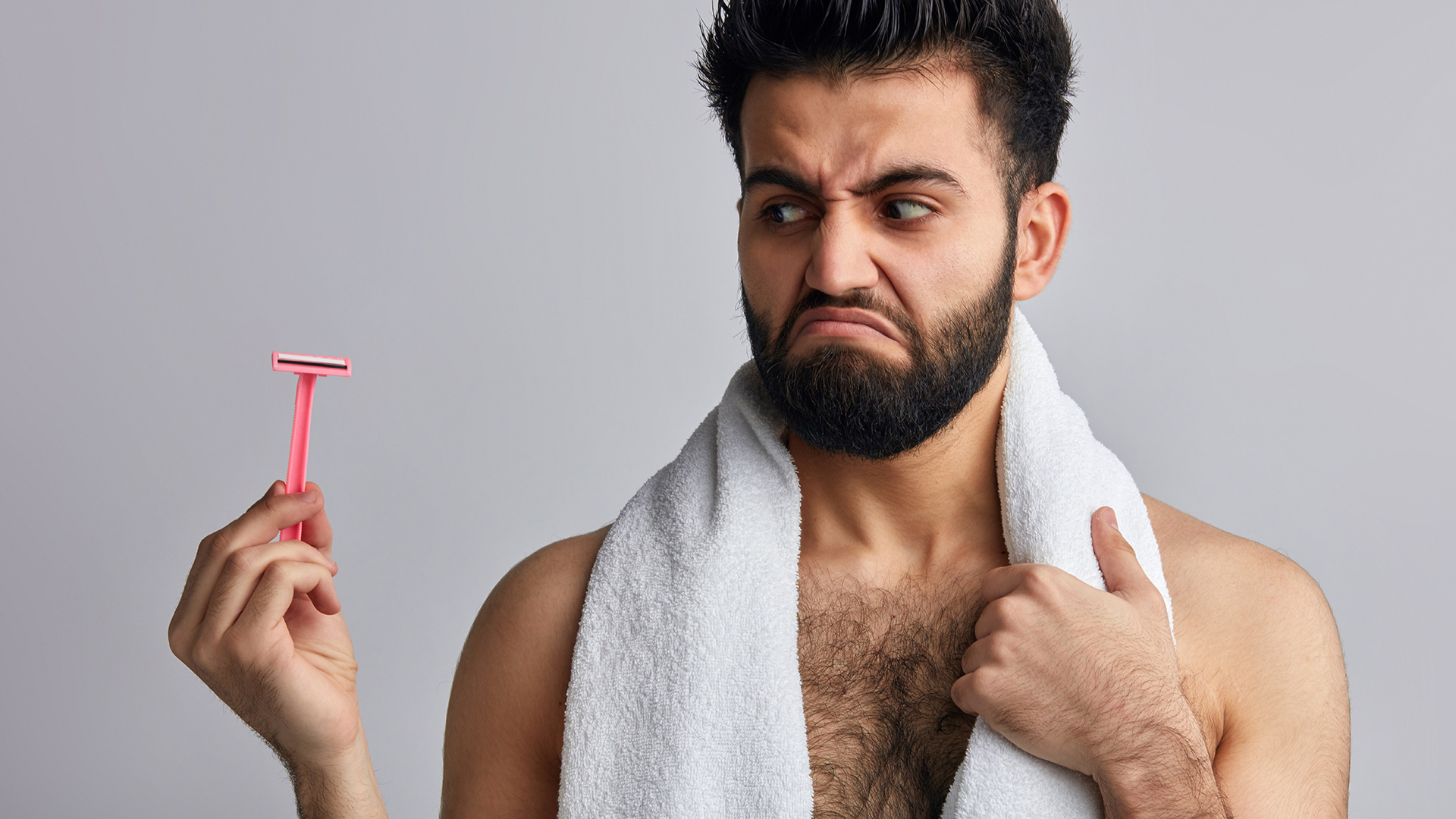 Brunette man looking like he just came out of bath with white towel around his nape, holding a pink razer and looking angry.