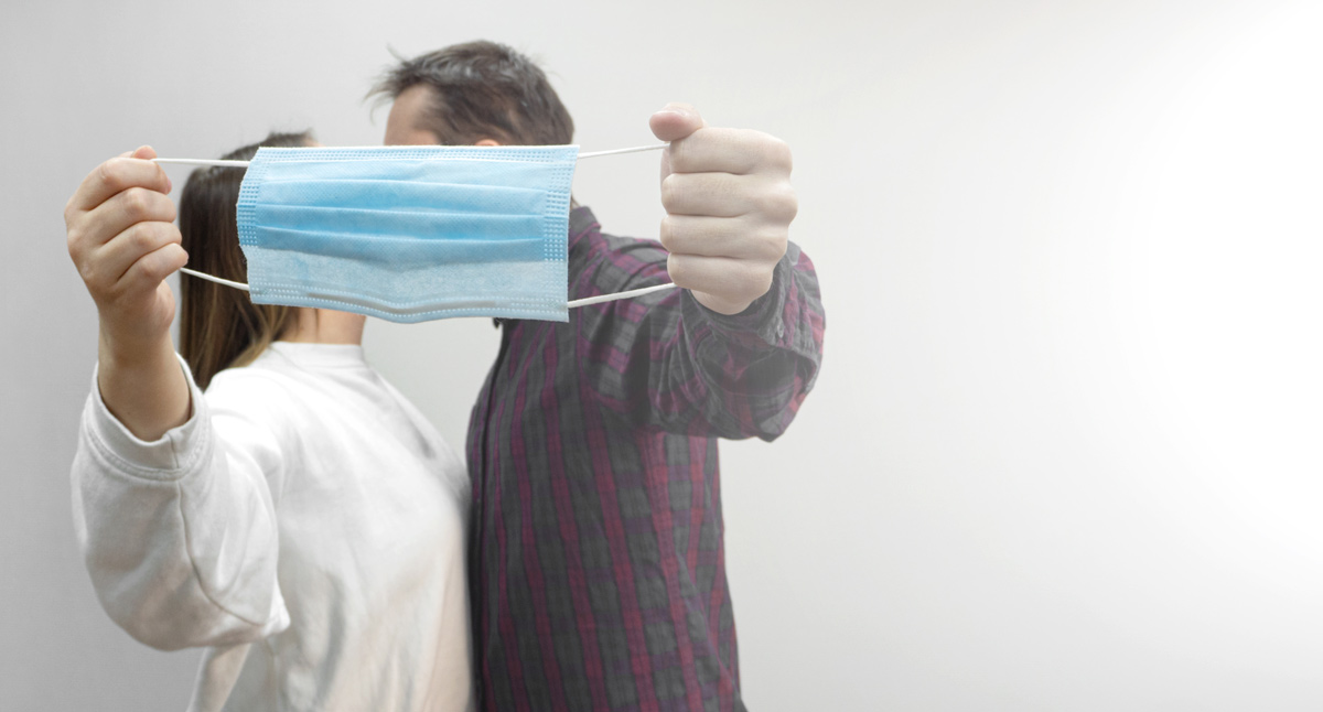 A man with a checkered shirt and a woman in a white jumper holding a facemask up to hide them from kissing 