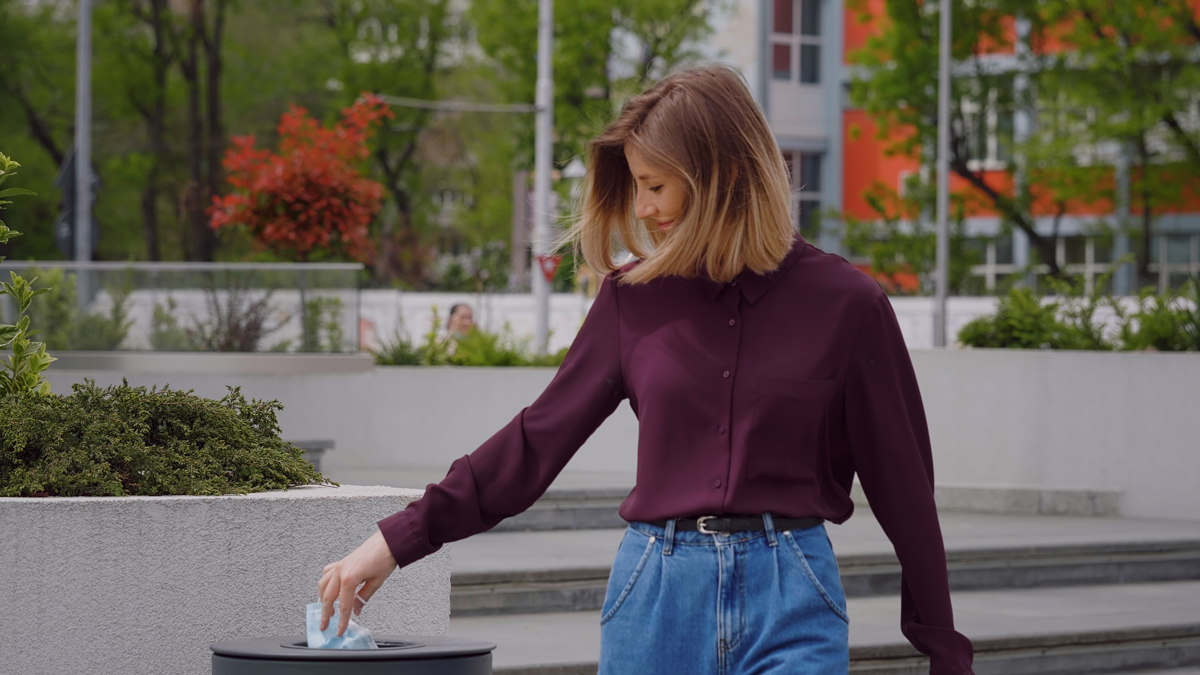 A woman walking through a park with a purple jumper and denim jeans putting a face mask in the bin