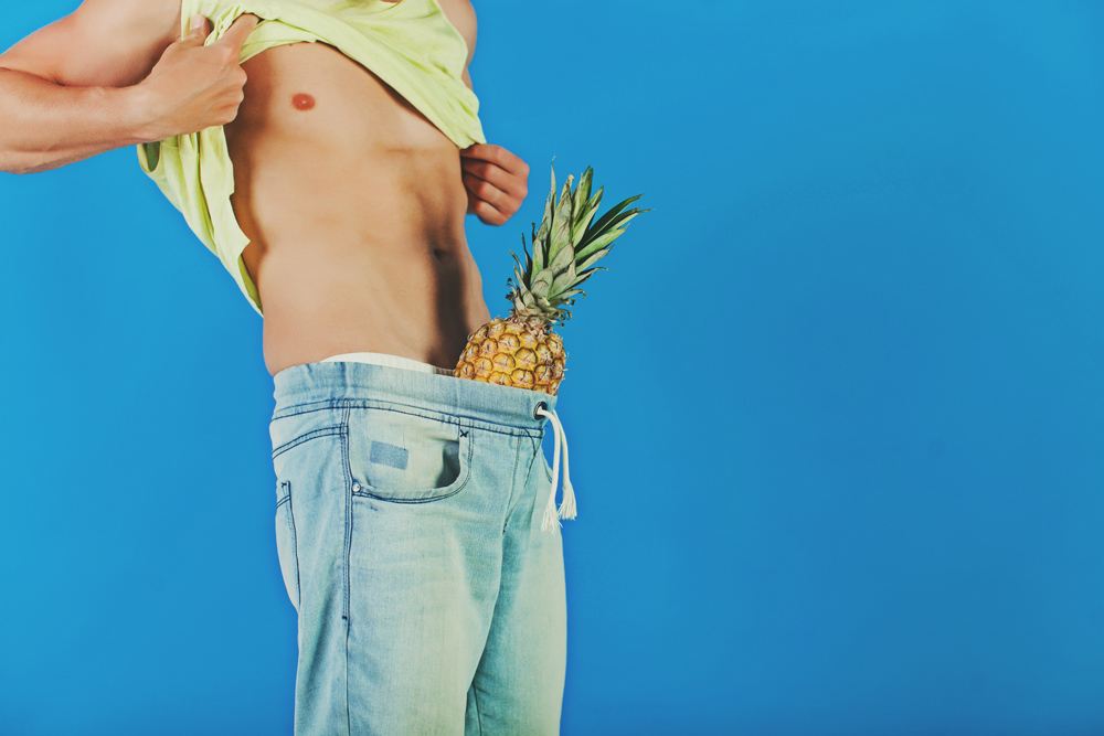 Man with yellow vest pulled up and denim jeans on, with a pineapple stuffed down the jeans.