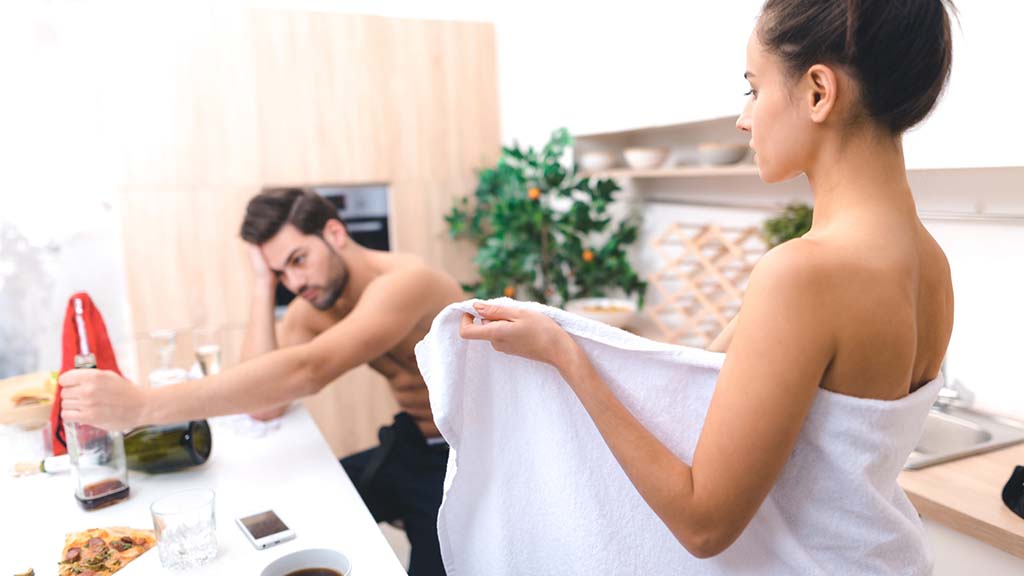 Man reaching for a bottle while a woman is standing diagonally from where he is and holding a white towel open towards him.