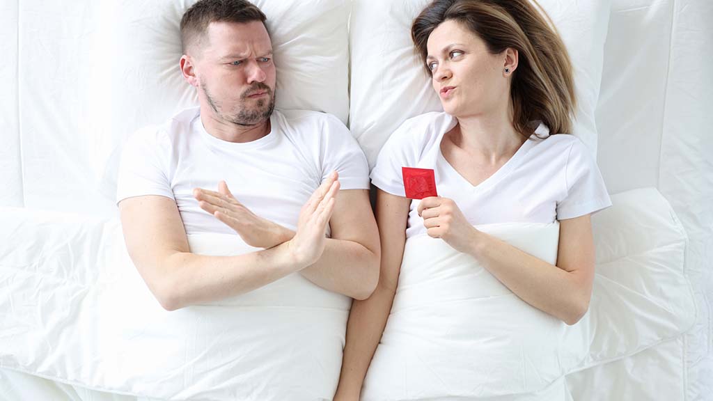 Blonde man and woman lying in a bed. The woman is giving the man a condom while he holds his hand to stop her.