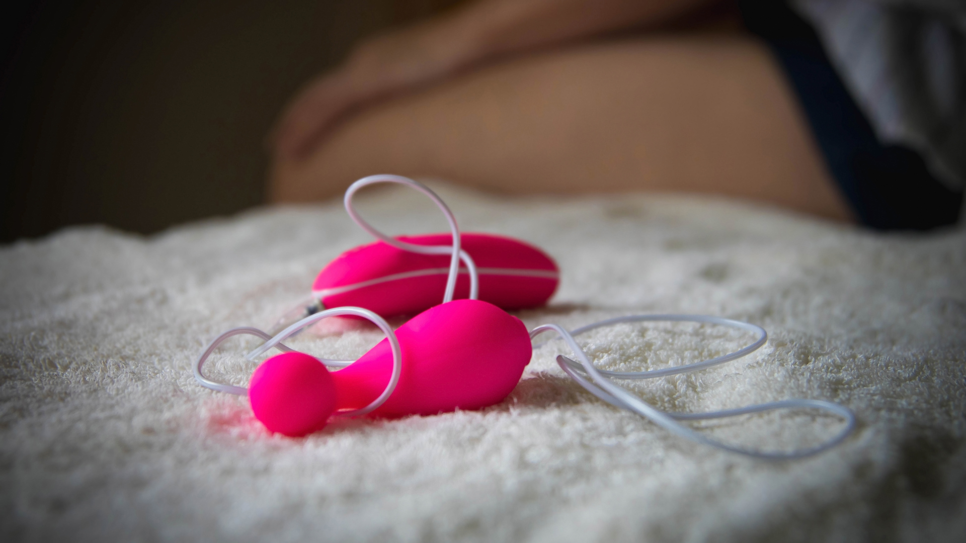 Pink egg vibrator with a wire on a white blanket, with a woman sitting on blanket in the background.