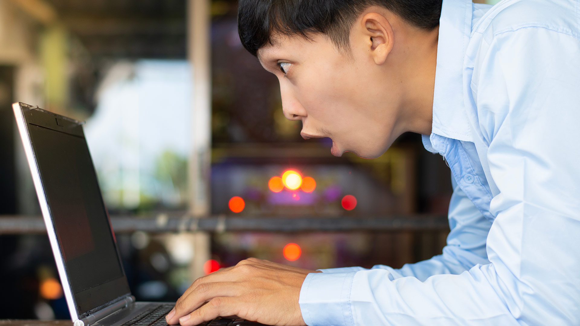 Brunette man in blue short standing up and looking shocked in front of his laptop screen.