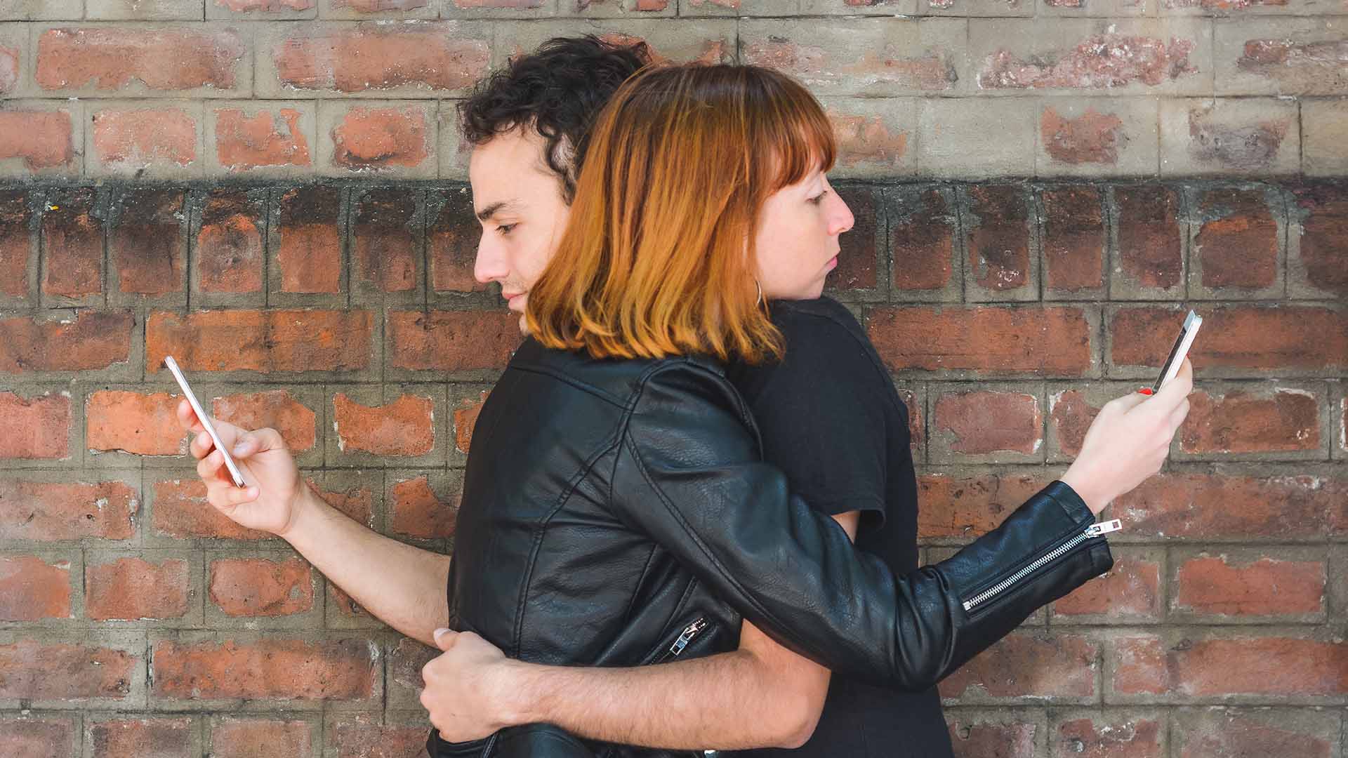 Orange-haired woman and brunette man wearing black, hugging each other while looking at their phones with one hand.