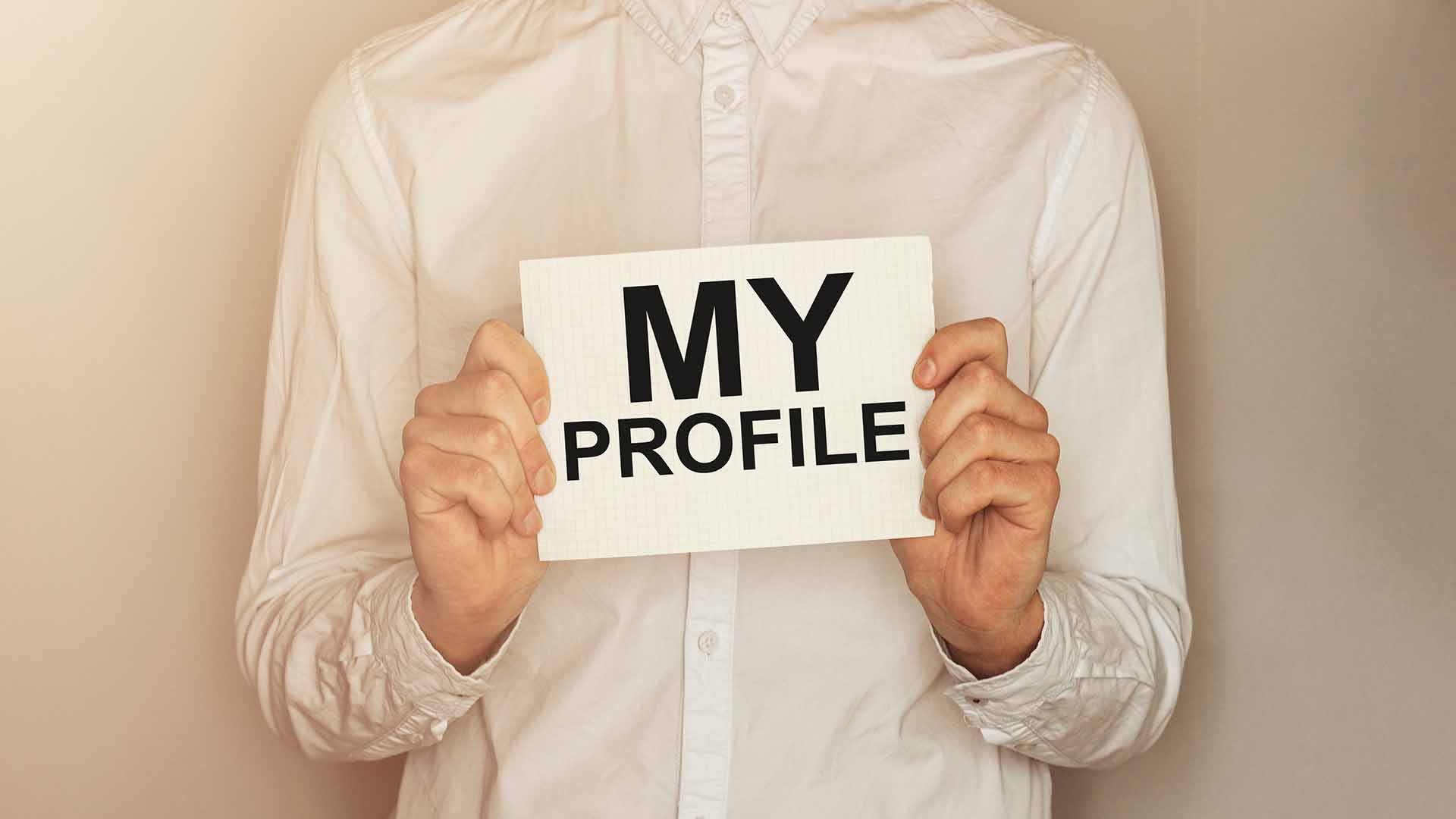 Torso of a man wearing a white shirt and holding a card reading "my profile" on it. Beige background.