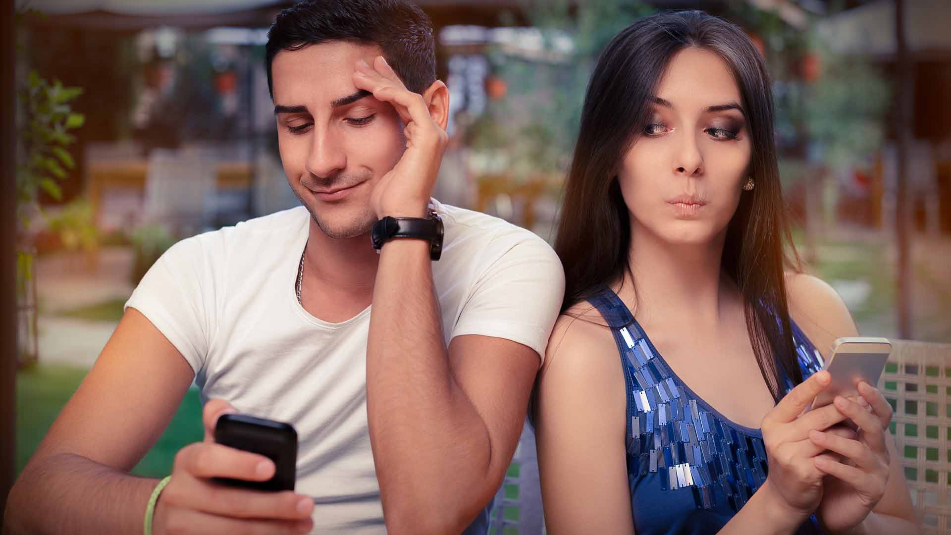 Brunette man in white shirt and brunette woman in blue shirt, sitting back to back and holding their phones.