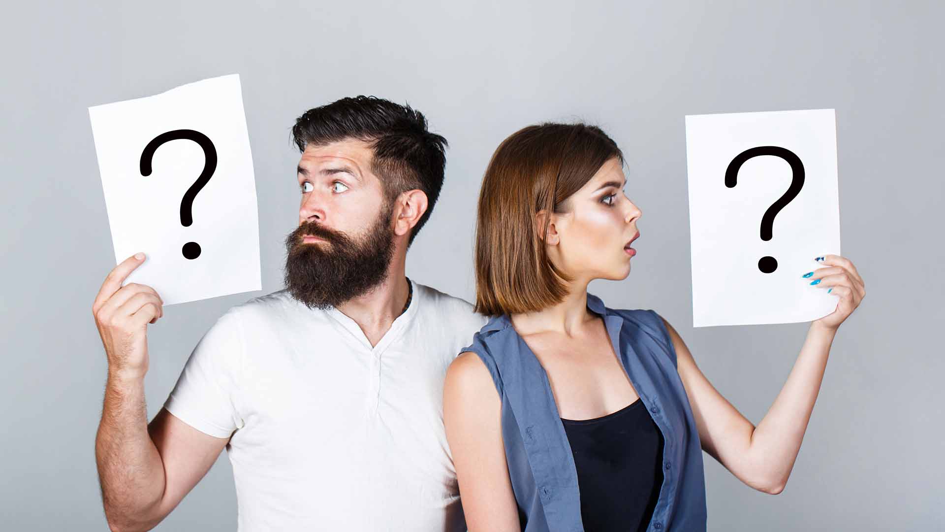 Brunette man in beard and white shirt standing next to woman in blue coat, both holding cards with question marks on them.