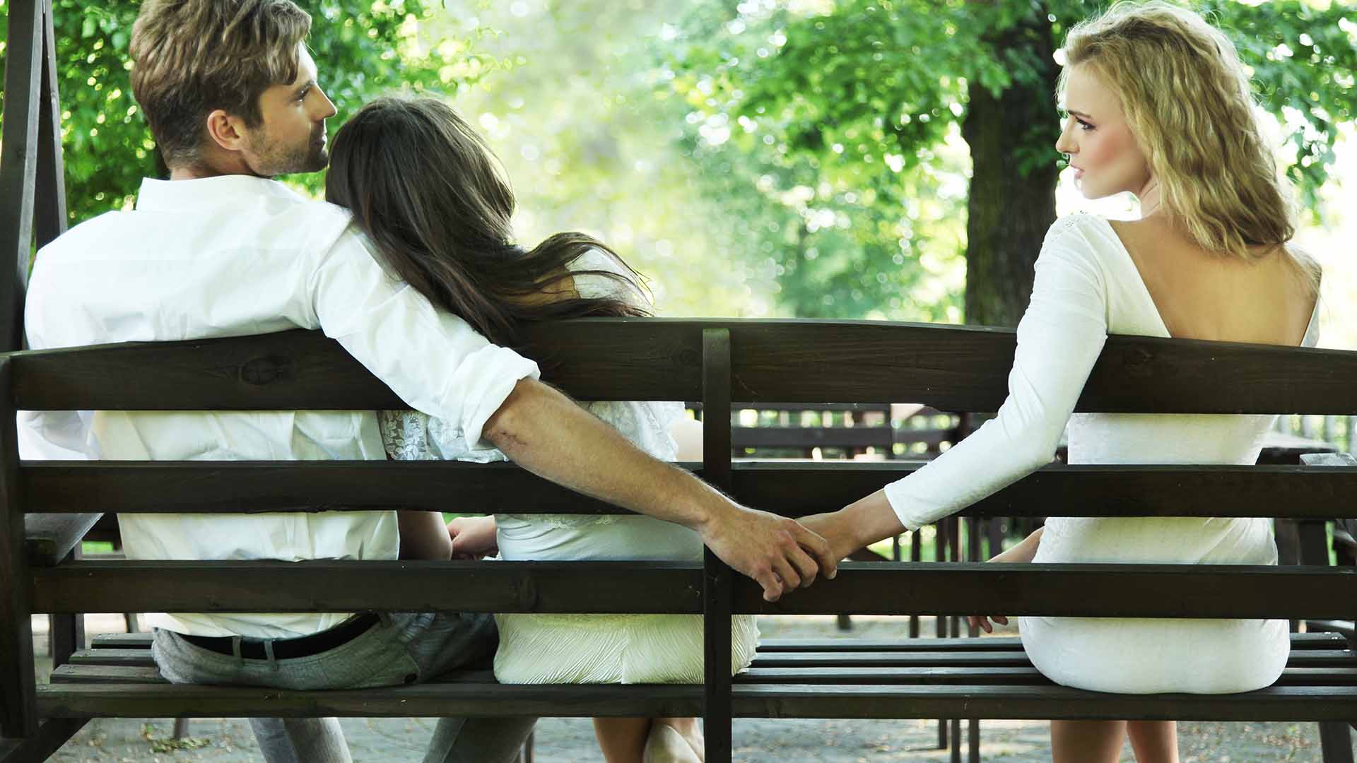 Brunette man and blonde woman sitting on bench at a park, holding hands behind brunette woman resting head on man's shoulder.