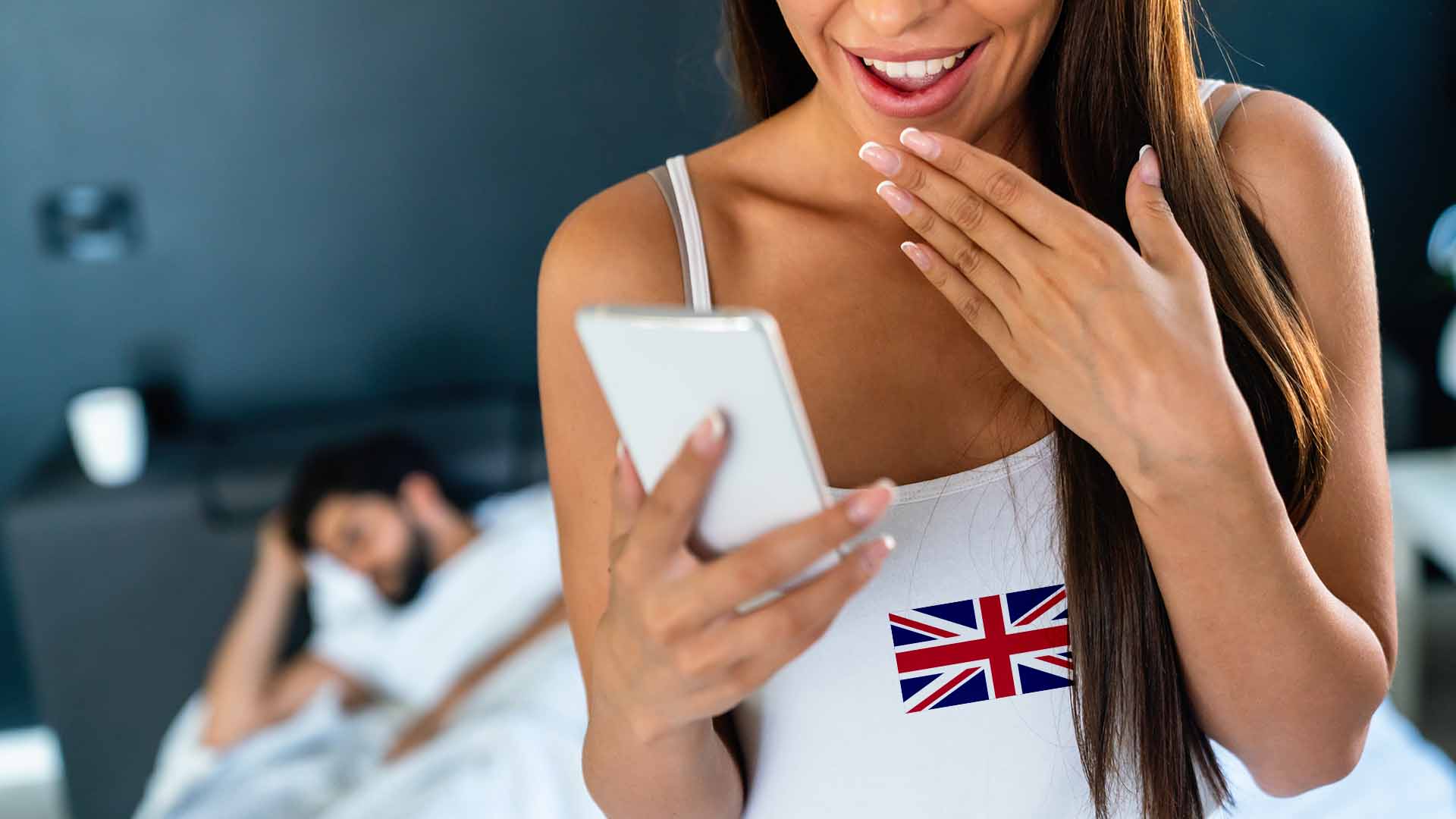 Brunette woman in white shirt with UK flag on it, holding her phone and smiling with brunette man sleeping in bed behind her.