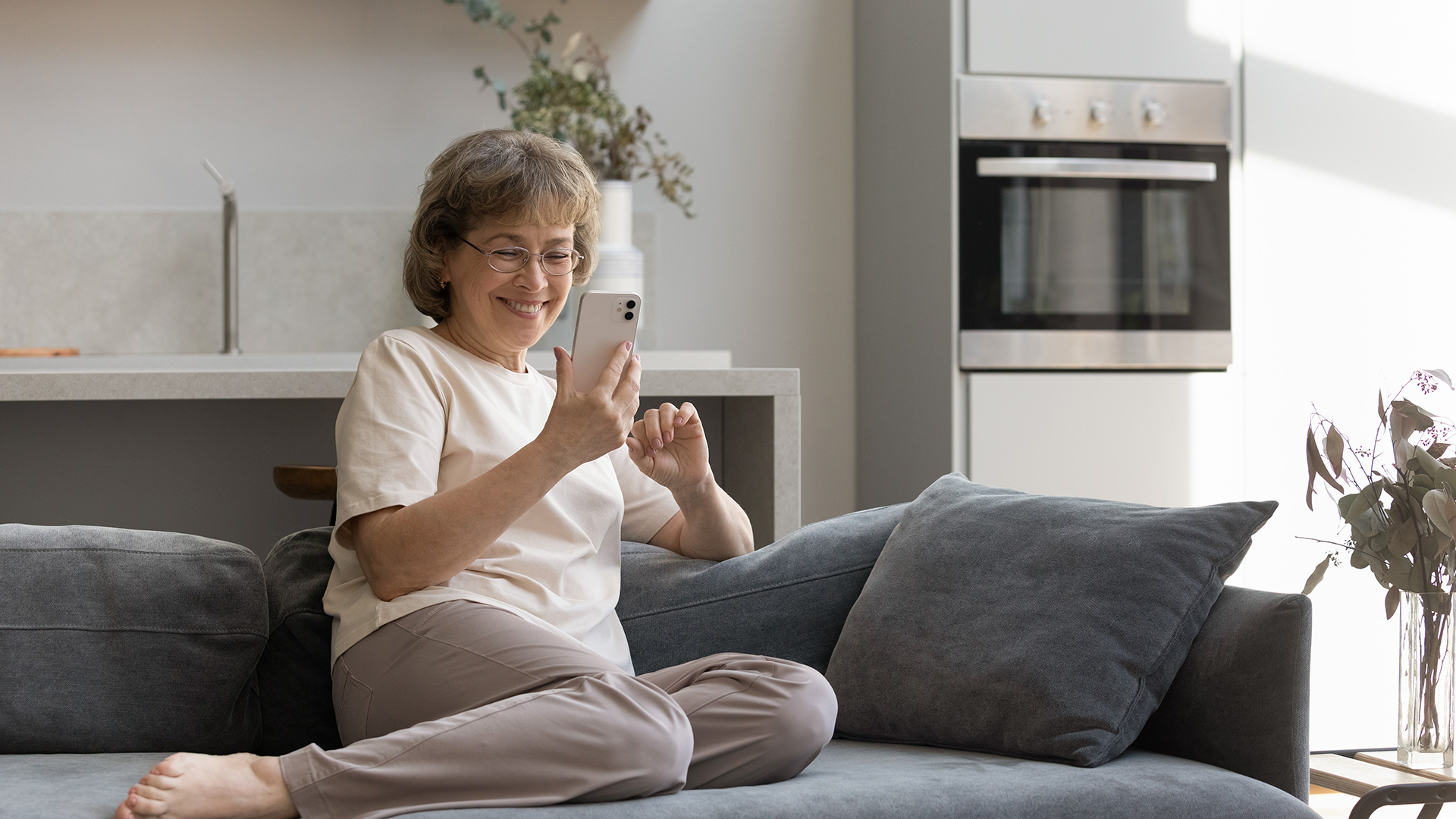 Older woman with grey hair, holding her mobile phone and smiling while looking at the screen. She is sitting on a grey couch.