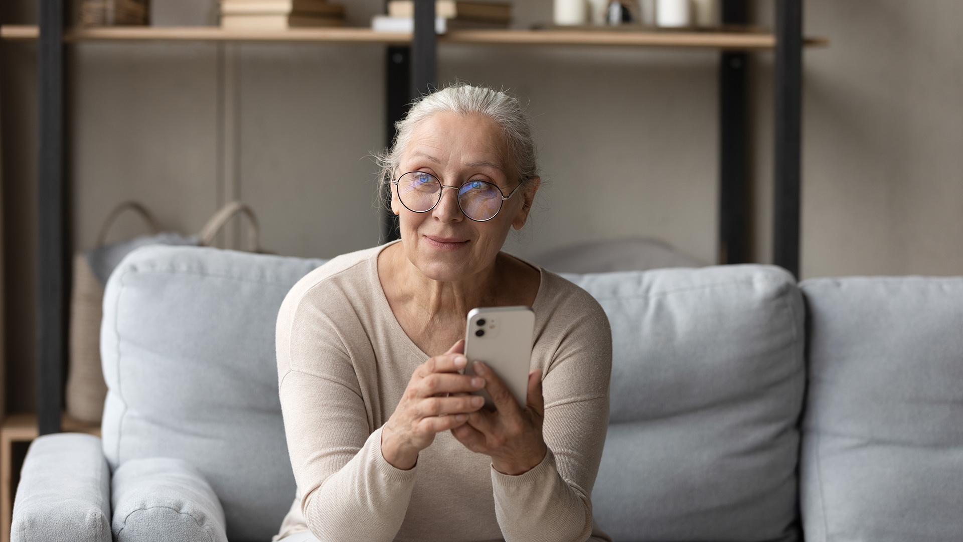 White-haired woman with glasses and beige t-shirt, holding her phone while sitting down on a couch. She is half-smiling.