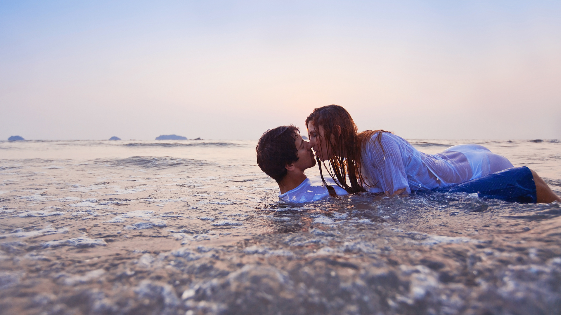 Woman in the sea wearing a see-through white dress pushing a man in an equally see-through white shirt in to the water.