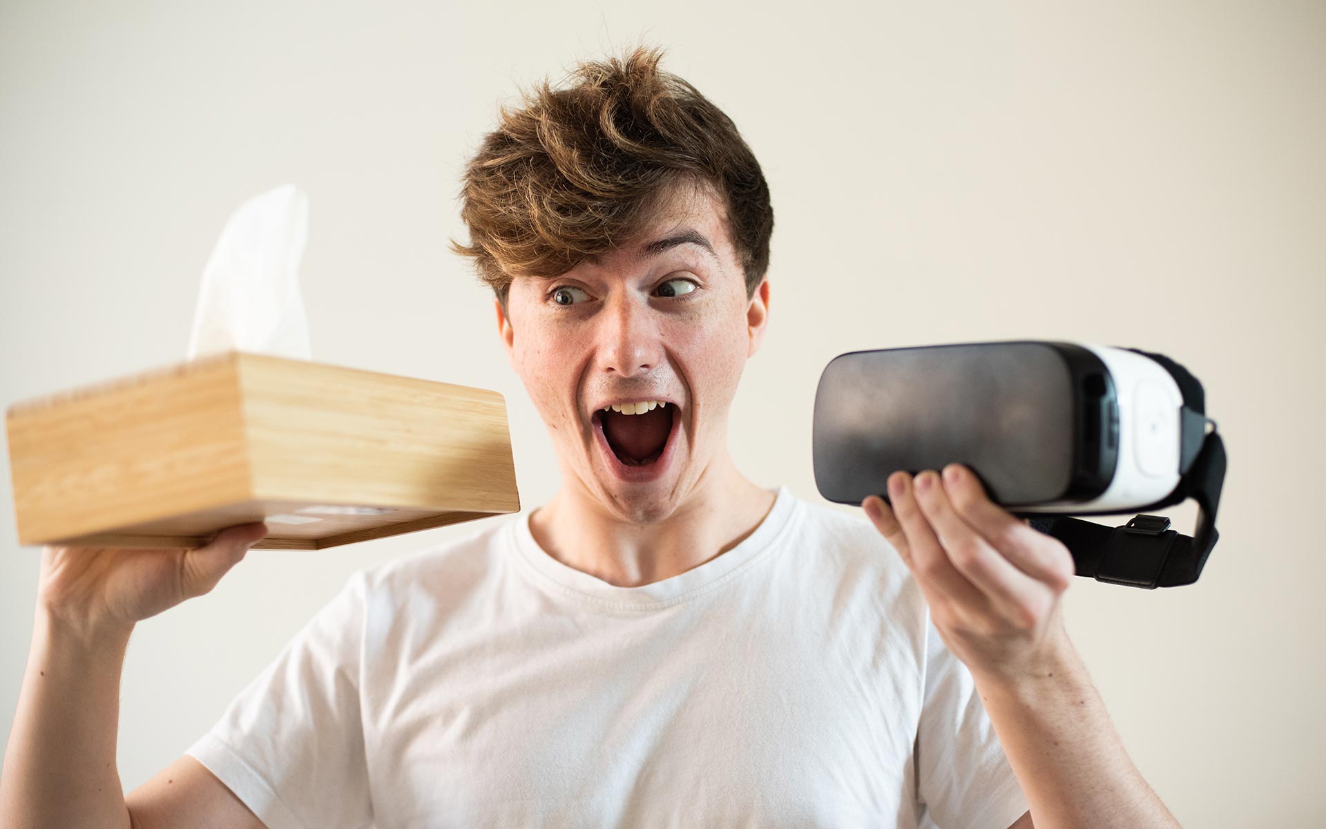 Brunette man in white shirt looking excited, holding a VR headset on one hand and a box of tissues on the other.