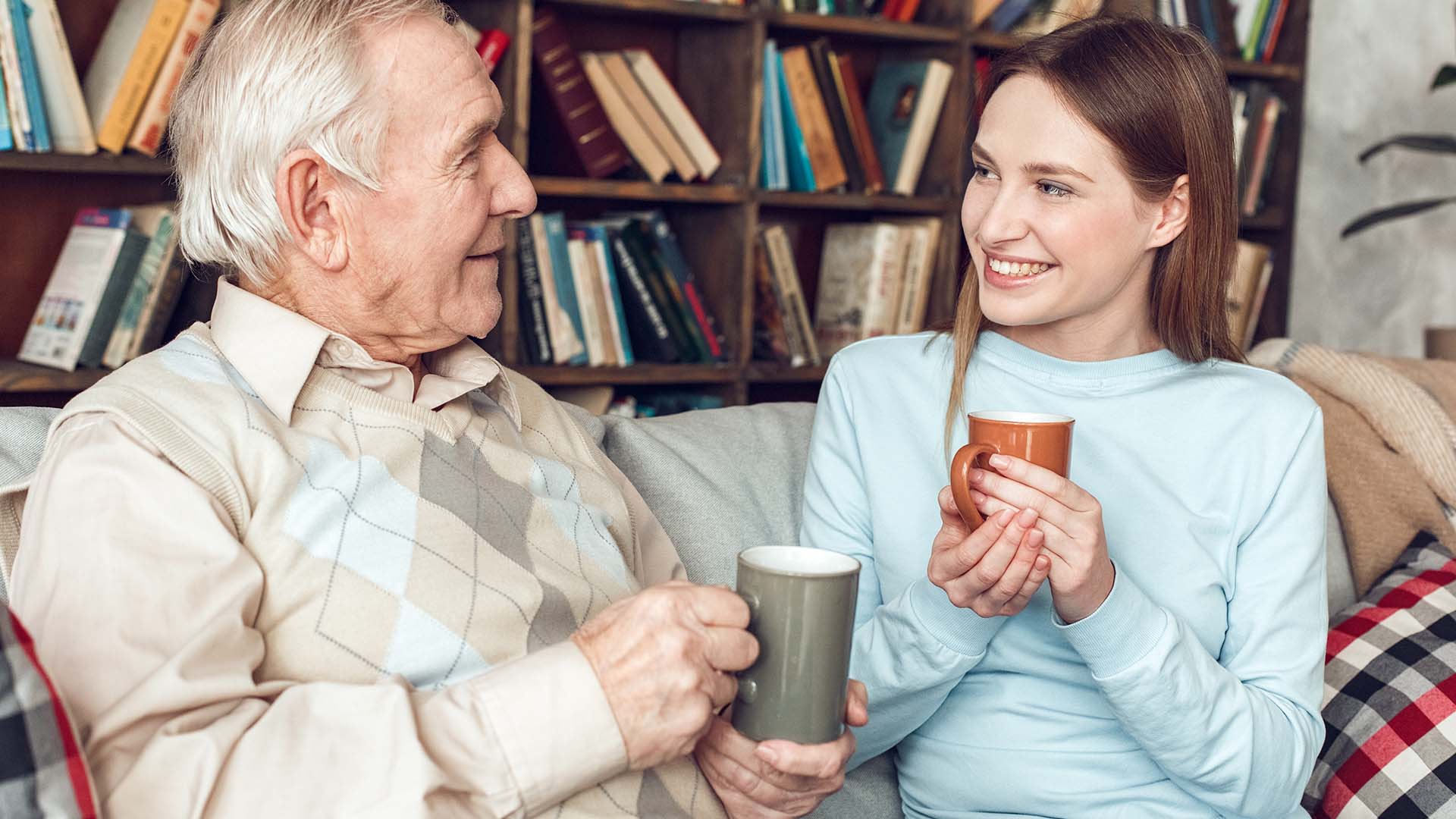 Older man and younger brunette woman sitting next to each other on a couch, conversing while holding a mug. Books behind them