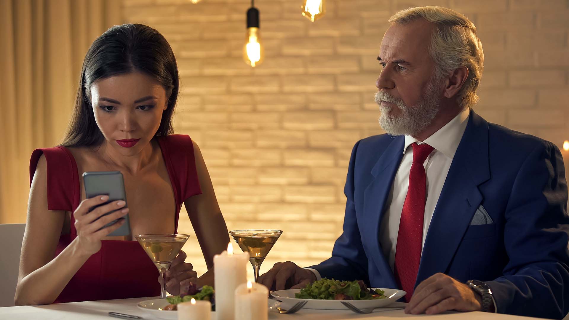 Older man in blue suit and younger woman in red dress having dinner. The woman is looking intensely at her phone