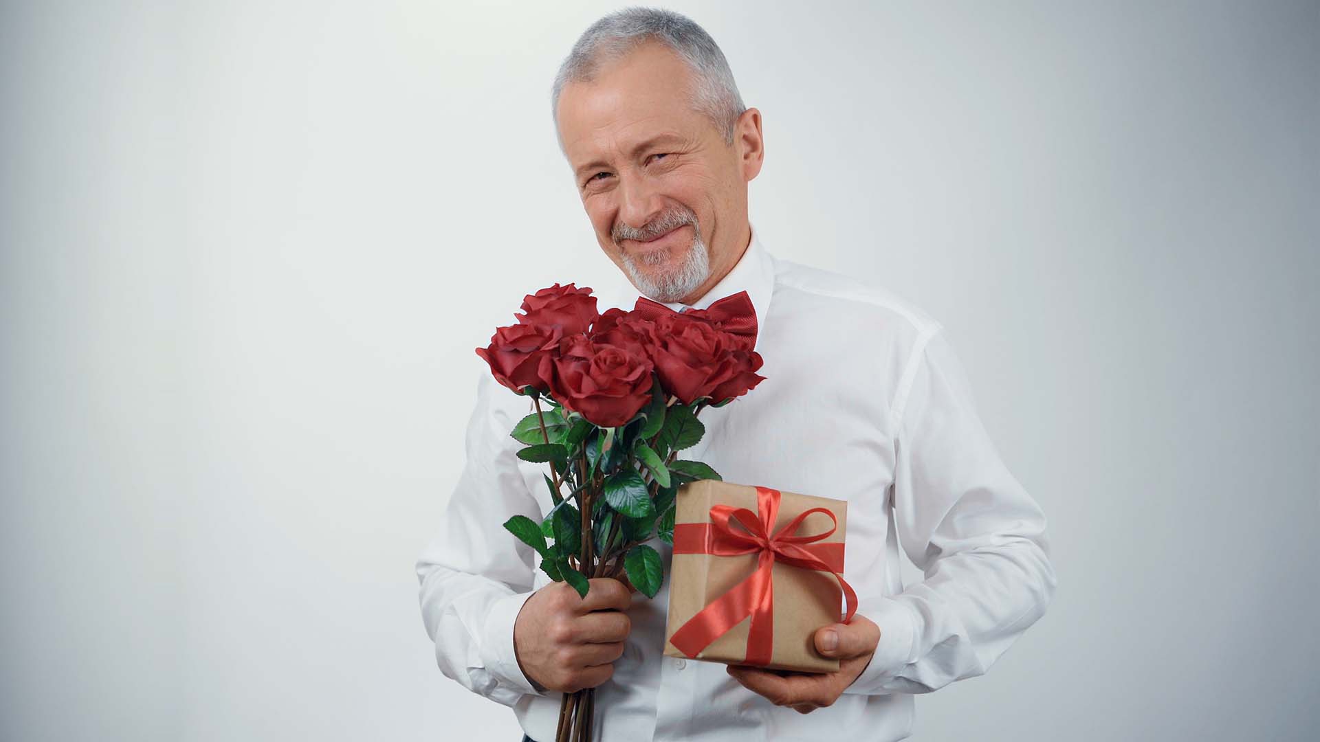 Older man in white t-shirt holding red roses and gift with red bow on it. Grey background.