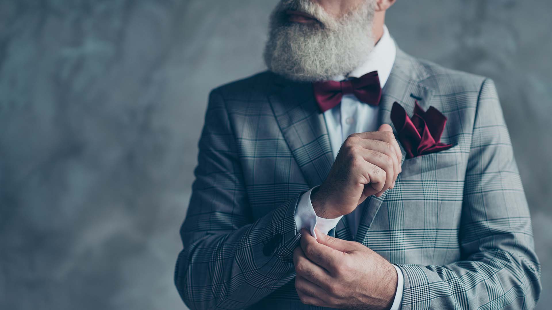 Older bearded man in grey checkered suit and red bow tie, fixing his sleeves while looking to the left. Grey background.