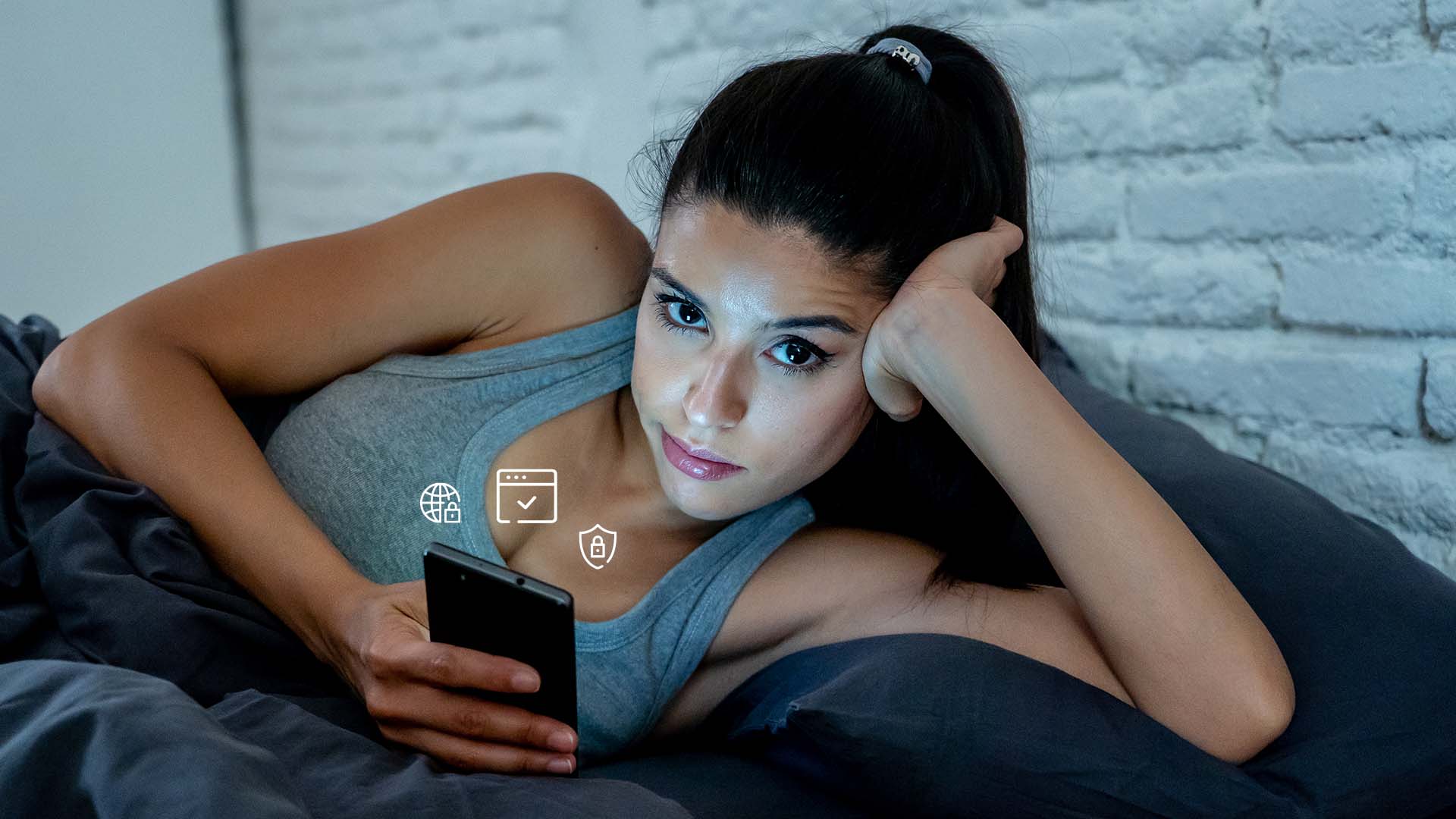 Brunette woman looking unamused while lying in her bed looking at her phone. Brick wall behind her.