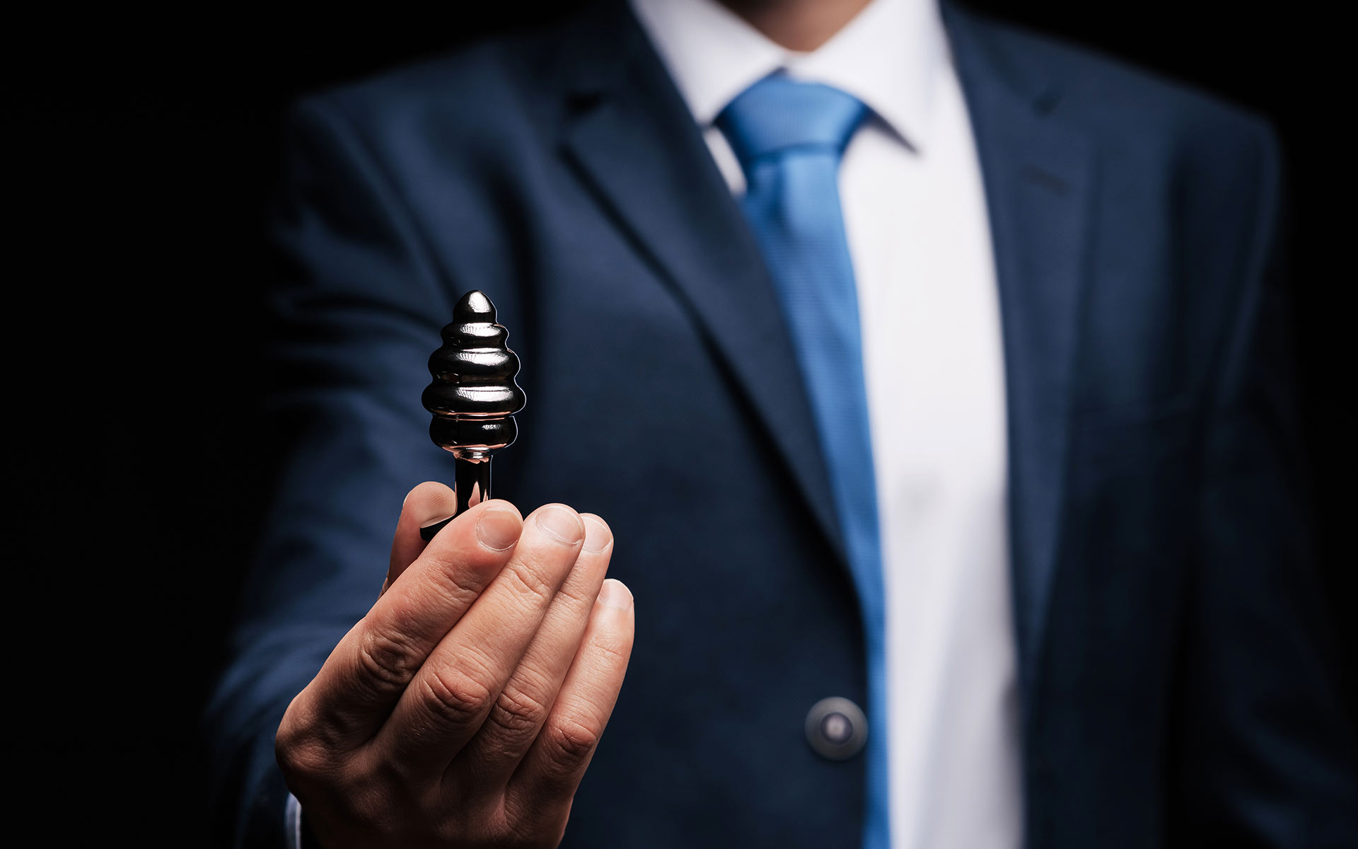 Torso of a man in a blue suit holding a stainless steal butt plug in front of him.