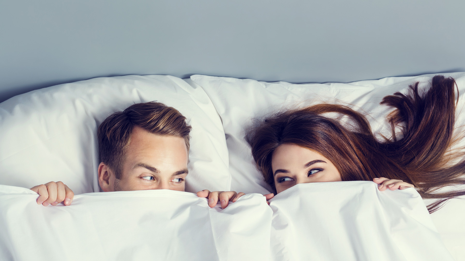 Blonde man and woman looking at each other and smiling while lying in bed of white sheets covering half of their faces.