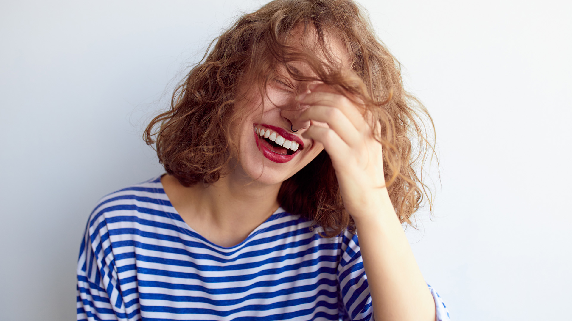 Brunette woman in red lipstick and blue-white striped shirt, holding the bridge of her nose while laughing.