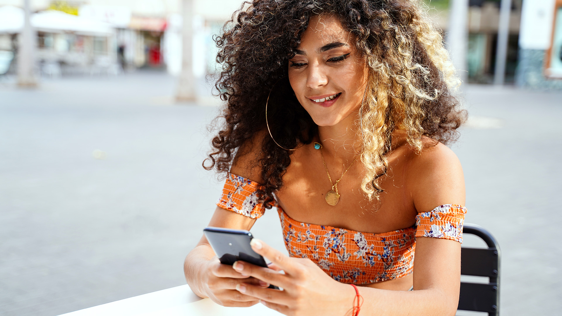 Curly-haired brunette woman sitting on a table in public and biting her lower lip while looking at her phone.