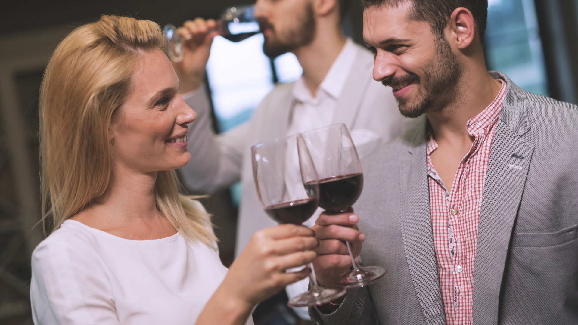 Blonde woman and brunette man making a toast while smiling. Man in the background drinking from his glass.