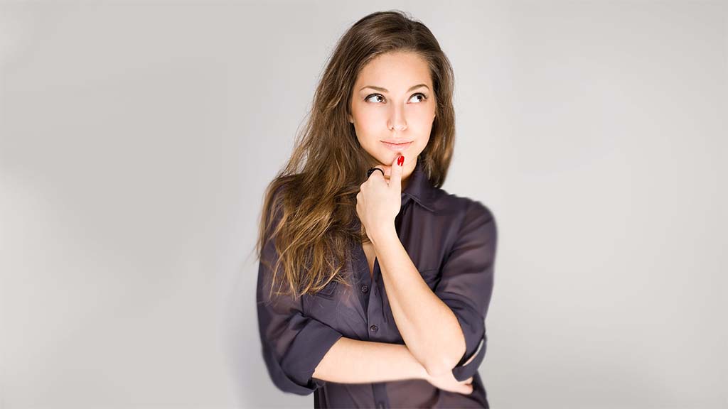 Brunette woman in dark purple short, holding her chin with her hand and looking upwards in question. Grey background.