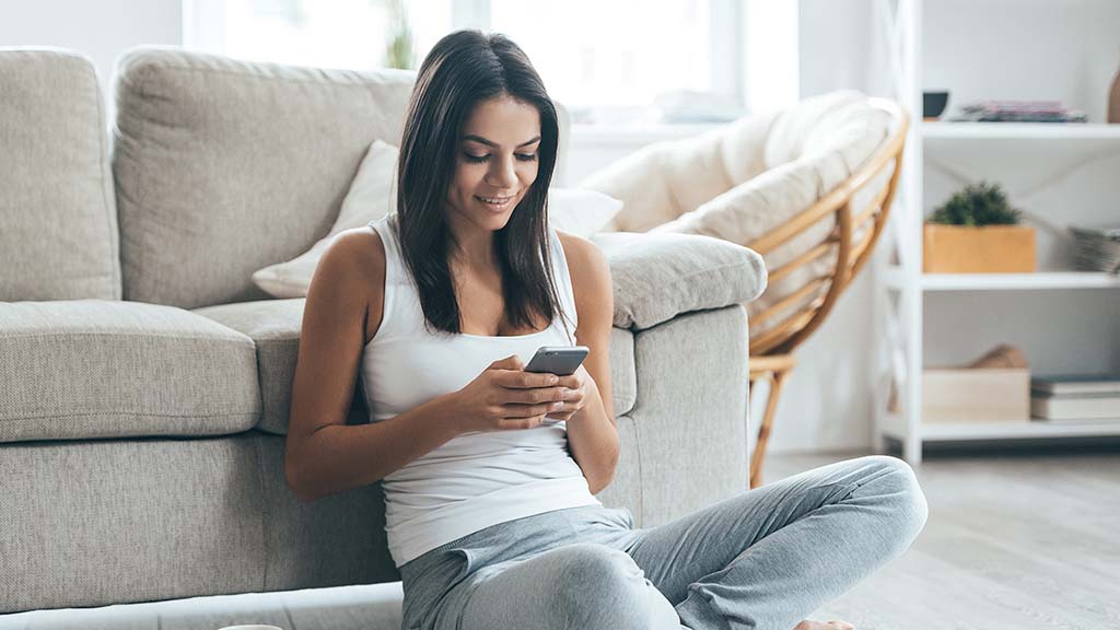 Brunette woman in white tank top, sitting on the floor in front of the couch, holding her mobile phone in her hands, smiling.