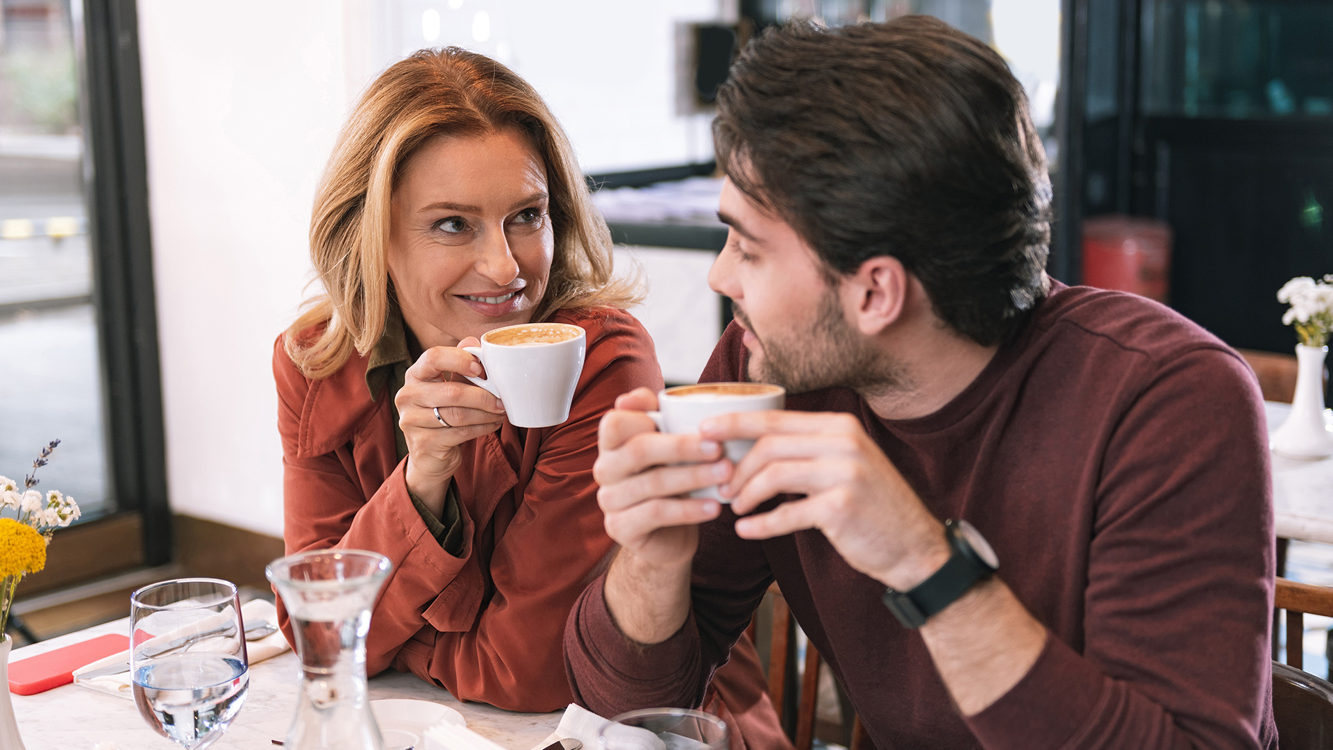 Blonde older woman in orange coat having a coffee date with brunette younger man wearing a brown shirt.