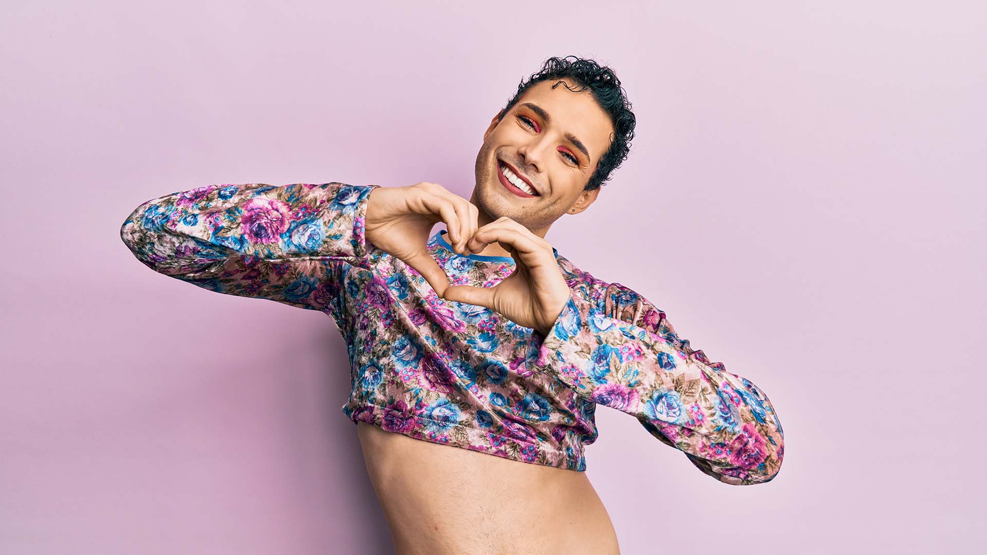 Dark haired trans person in colorful long-sleeved crop top, forming the shape of a love heart with their hands and smiling.