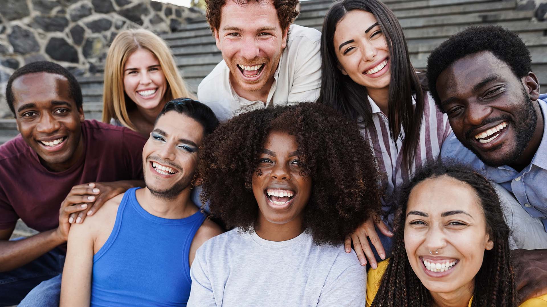 A diverse group of people sitting on stairs next to a wall of rocks, smiling and laughing together.