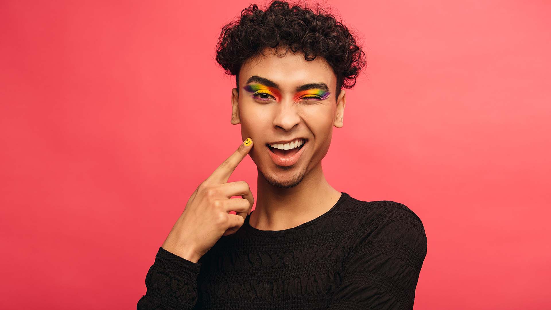 Brunette trans person in rainbow eye shadow in front of red background wearing back shirt, winking while smiling.