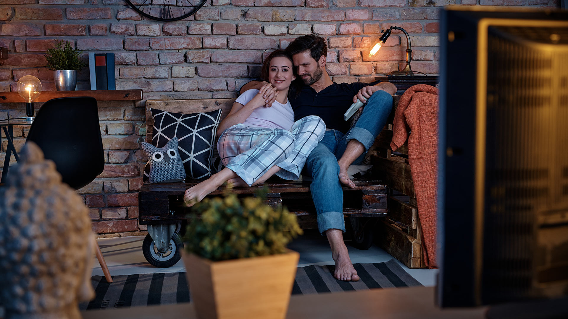 Brunette man and woman cuddling on a couch and watching TV. The man has one hand around the woman's body, holding her hand.