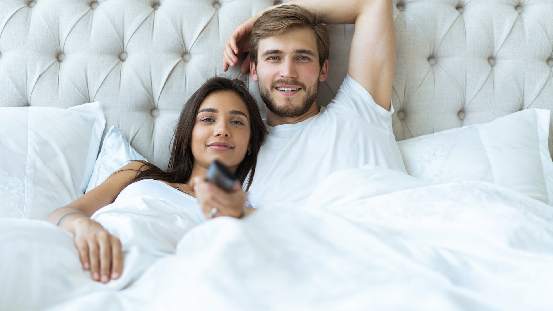 Brunette man and woman both in white t-shirts lying in bed next to each other. The woman is holding a TV controller.